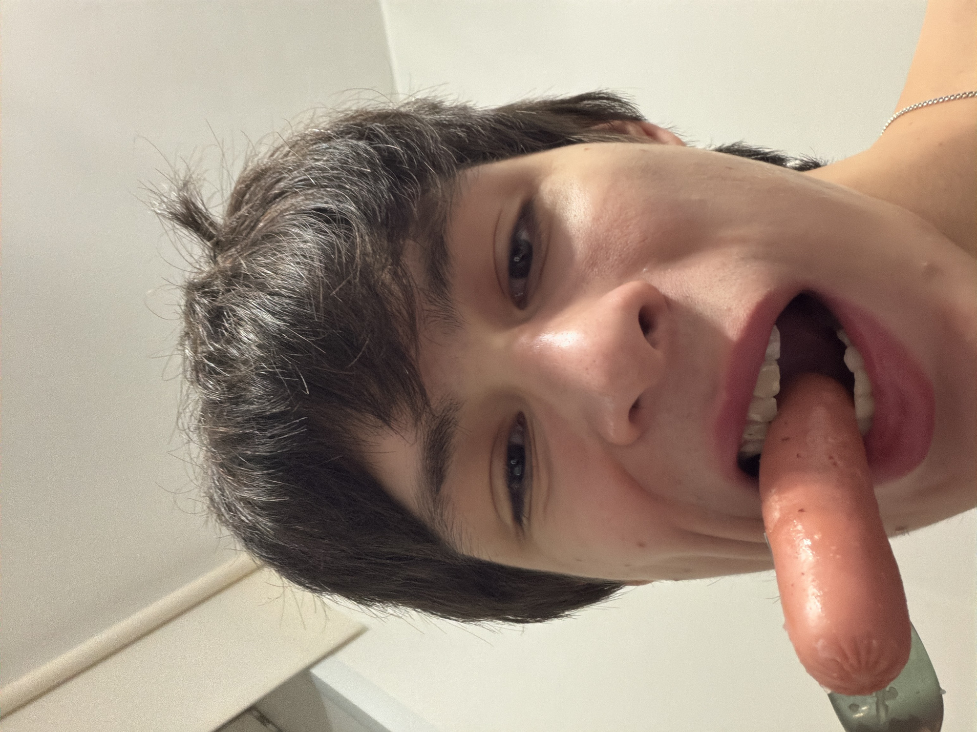 The image presents a close-up, head-and-shoulders view of a young individual with medium-dark brown, disheveled hair, standing indoors in the city of Yekaterinburg, Russia. The individual, appearing to be male, looks directly at the camera with a subtle expression. Their mouth is open wide, partially consuming a pinkish hot dog held by a silver fork. The tines of the fork are visible beneath the hot dog as it enters the mouth. Some moisture is apparent on the fork. A thin silver chain is visible on the individual's right collarbone. The background consists of plain, light-colored walls and ceiling, suggesting an interior space with even illumination.