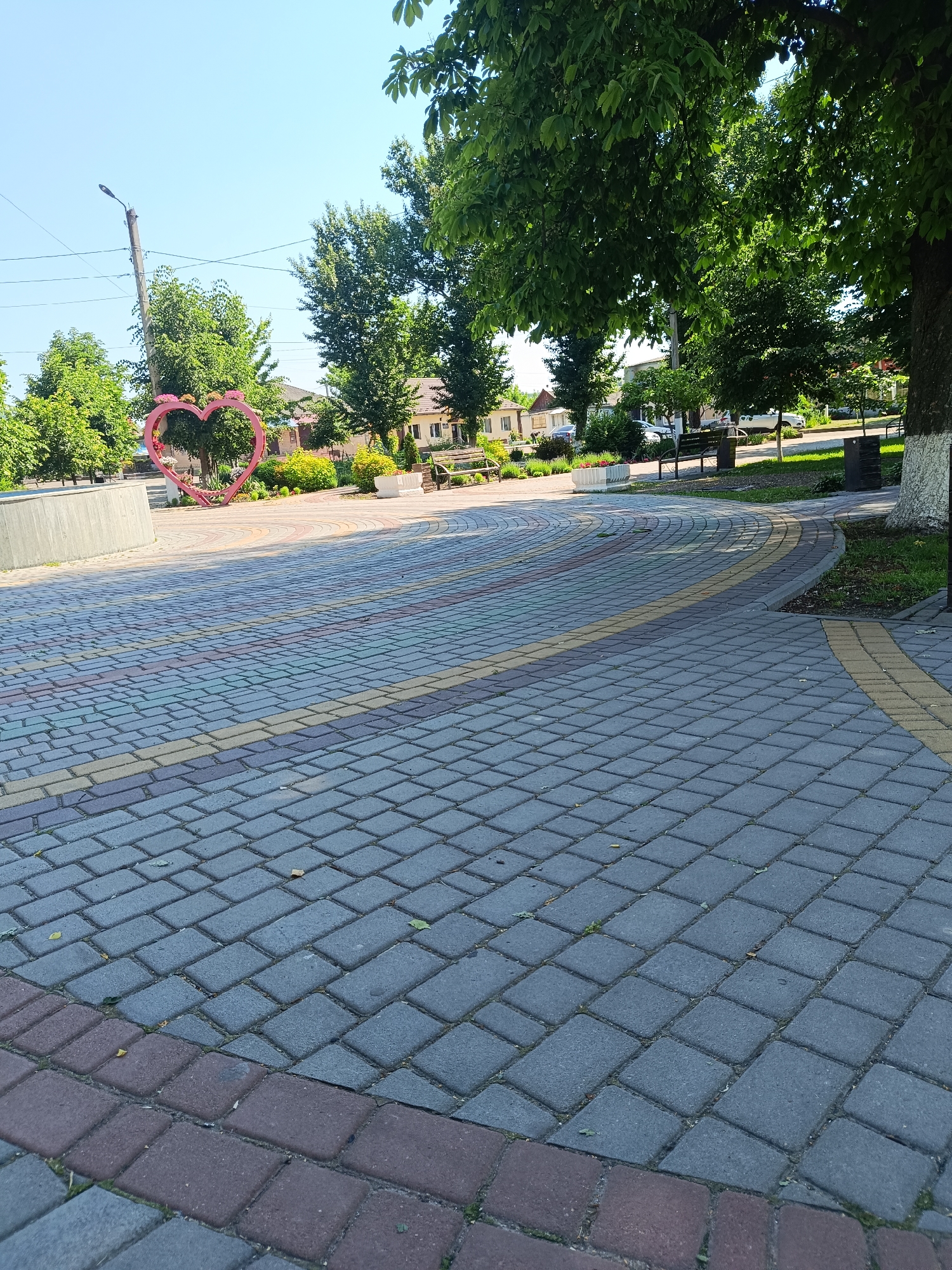The image depicts a paved plaza in Okny, Ukraine, featuring a large, pink, heart-shaped sculpture adorned with flowers in the background. The plaza itself is paved with interlocking bricks in shades of gray, yellow, and red, forming a circular pattern that recedes into the distance. Surrounding the plaza are lush green trees and some buildings. A concrete structure is visible on the left, and benches and trash receptacles are placed around the area. The sky is clear and blue, indicating a sunny day. The perspective is from ground level, looking across the plaza towards the heart sculpture and the surrounding environment. No people are present in the image.
