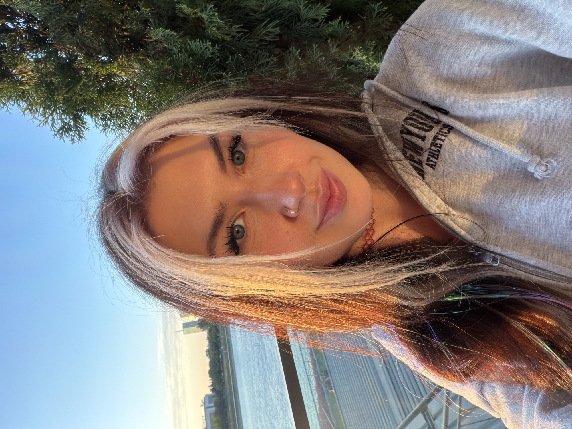 A young woman is captured in a rotated selfie, positioned from the chest up, with her head gently tilted. She has long, multi-toned hair, featuring prominent blonde highlights that frame her face and blend into darker, reddish-brown strands. Subtle iridescent tinsel or hair extensions in shades of green and purple are visible within her darker hair. Her eyes are a striking green, and she wears a silver hoop piercing in her left nostril. A soft, pleasant smile graces her face as she looks directly into the camera. She is dressed in a light grey hooded sweatshirt, with the text "NEW YORK ATHLETICS" printed in a dark font visible on the upper chest/shoulder area.
The scene is set outdoors during what appears to be the late afternoon or "golden hour," as indicated by the warm, natural light illuminating her features and hair. In the background, a clear blue sky with a few faint clouds transitions into a dense canopy of dark green, coniferous trees. Beyond these, a wide body of water, likely a river, stretches out, reflecting the soft light. Indistinct buildings are visible on the distant bank, and a dark railing or fence structure is partially seen in the foreground, separating the woman from the water. Given the context, this scenic view is situated in Belgrade, Serbia, possibly along one of its major rivers.