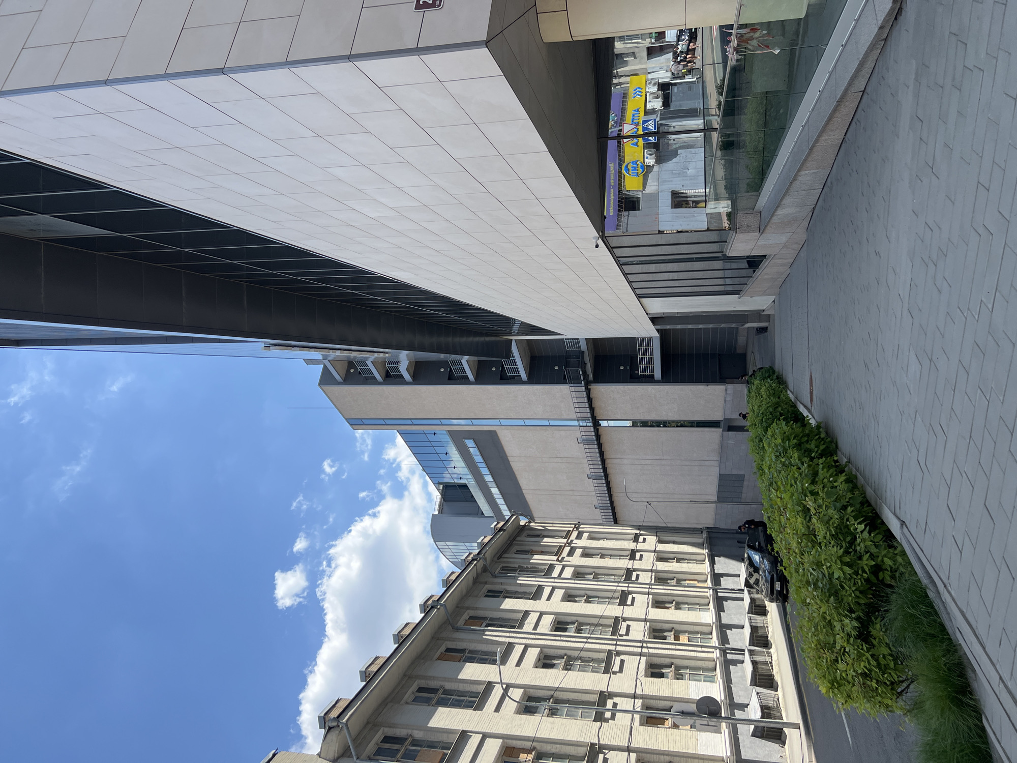 This image depicts a street scene in Kharkiv, Ukraine, featuring a modern glass-fronted building adjacent to an older, lighter-colored building with multiple windows and visible chimneys. A dark sedan is parked on the street next to a row of lush green bushes. A person in what appears to be a uniform stands near the car. The street is paved, and a sidewalk with patterned gray tiles leads away from the viewer. The sky above is blue with scattered white clouds. In the background, through the lower levels of the modern building, signs with Ukrainian text and logos are visible, including one with a yellow background and blue lettering.