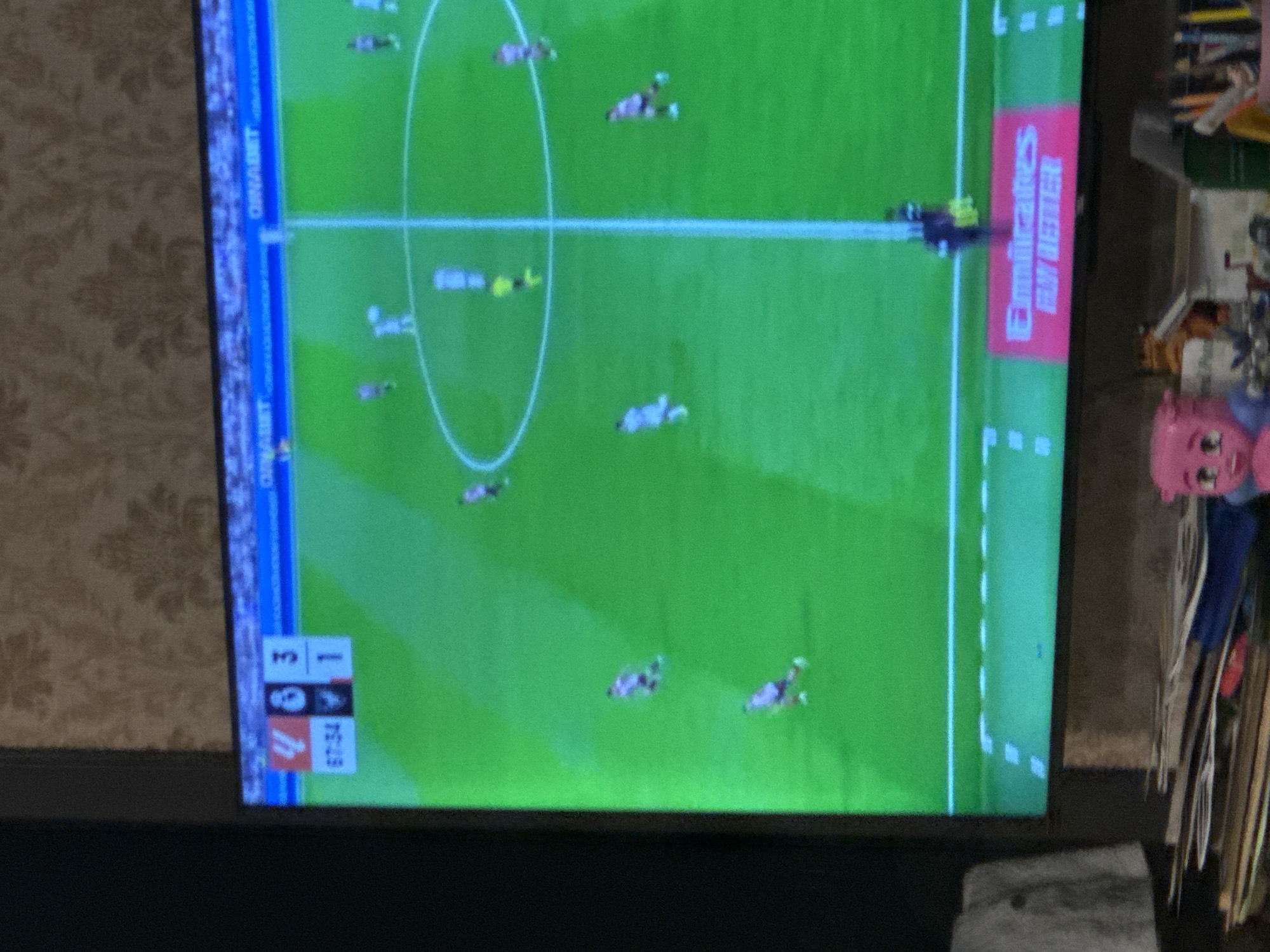 The image shows a television screen displaying an Australian Rules Football match. The scoreboard indicates a score of 3-1 (unknown teams). The "Emirates Fly Better" advertisement is visible on the field. It appears that the photo was taken indoors, possibly in Yerevan, Armenia, although there is no specific indication in the frame itself to verify its location. There are various miscellaneous objects surrounding the TV screen.