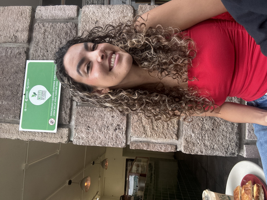 An adult female, estimated to be in her 20s-30s with long, curly, highlighted brown hair, is seated and smiling at the camera. She wears a red strapless top and blue jeans. To her left, on a light-colored table, an iced coffee in a white cup and three pieces of a grilled sandwich on a red plate are visible. Behind her is a textured stone wall or pillar. Mounted on this wall above her head is a rectangular green sign with white Spanish text, including "SALUD," "COFEPRIS," "CONADIC," and declaring the space "100% LIBRE DE HUMO Y EMISIONES" (100% Smoke and Emission Free Space), along with contact numbers for COFEPRIS and a "LÍNEA DE LA VIDA." These governmental entity references and language confirm the location in Mexico City, Mexico. To the left of the stone pillar, an interior space is visible, featuring a counter with display cases, dark green tiling, and two ceiling-mounted pendant lights. A person, partially obscured, is visible behind the counter within this interior space.