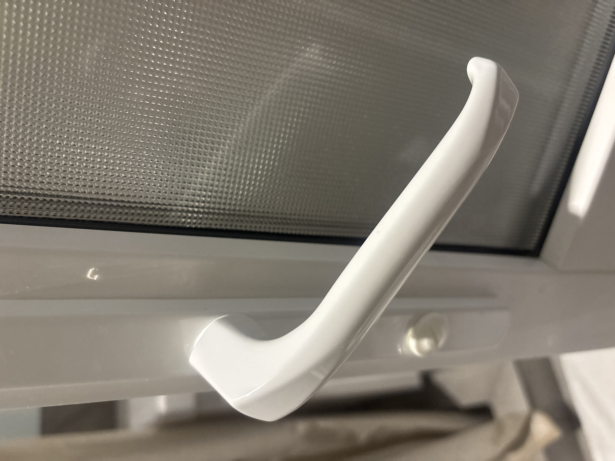 The image shows a white window handle, made of plastic, attached to a white window frame. The window frame appears to be part of a larger structure. Behind the handle and frame, a textured glass panel is visible, likely a window pane with a decorative or obscuring pattern. To the left of the window, a beige curtain is partially visible, suggesting the interior of a room. The lighting in the image appears to be artificial, with some highlights reflecting off the glossy surface of the handle and frame. The location is stated to be the city of Ldzaa, Abkhazia.