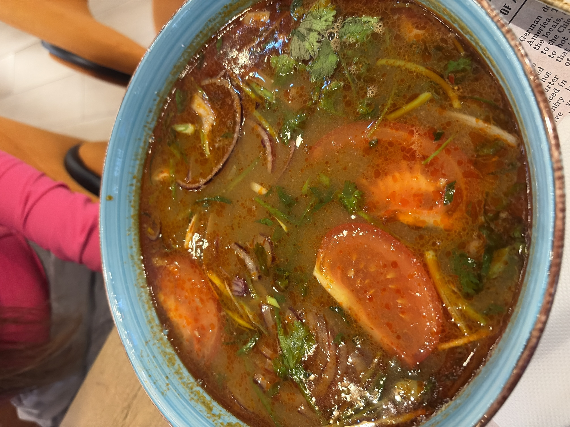 The central focus of the image is a vibrant bowl of hot soup, presented in a distinct blue bowl with a textured, striped rim. The soup itself is a rich, reddish-brown broth, featuring a glistening, oily surface indicative of a flavorful and possibly spicy preparation. Prominent ingredients visible in the soup include large slices of red tomato, fresh green herbs (likely cilantro), thin yellowish-white strands (possibly lemongrass or ginger), and slices of red onion. There are also smaller, lighter-colored pieces, which could be meat or seafood.
In the background, a person wearing a bright pink long-sleeved top is partially visible on the left, along with blurred brown and black furniture, possibly a chair or booth, and a light-colored floor or wall that appears to be tiled. On the right side, a crumpled piece of newspaper or print material is visible next to the bowl.
The visible text fragments from the newspaper include words such as "OF," "German do," "America," "the locals," "hot," "to the Chic," "that," "e," "ed," "arter," "ced in," "ntry i," and "ve yo." These suggest content related to a news article, possibly discussing German history or people in America, and may mention something "hot." The scene appears to be set indoors, likely in a restaurant or home dining setting, with overhead lighting creating reflections on the soup's surface. The overall impression is a close-up view of a ready-to-eat meal.