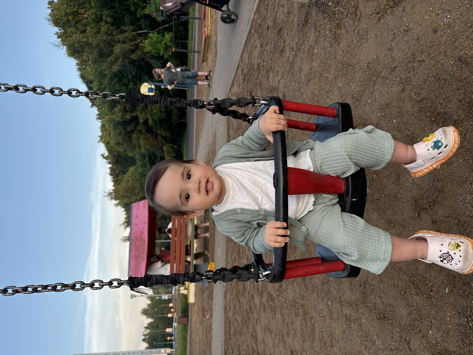 A toddler sits on a playground swing. The child is wearing a light green outfit and pink shoes. The swing is red with black and metal accents. The background shows trees, a building with a red roof, a basketball hoop, and other playground equipment. The ground is covered in sand. The sky is blue with some clouds. The image appears to be taken at a public park.