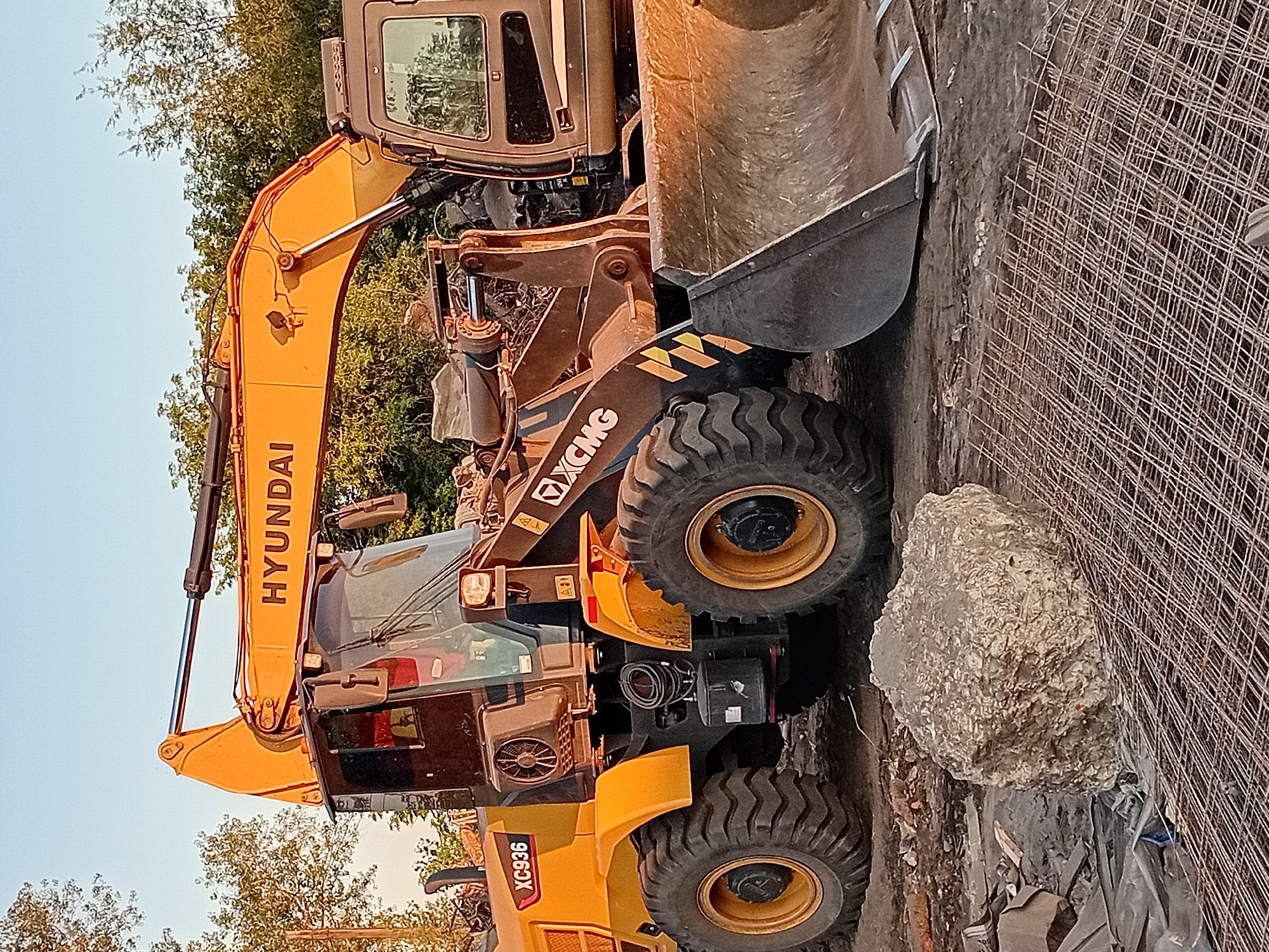 The image shows a construction site in the city of Semey, Kazakhstan, with heavy machinery present. In the foreground, a large, irregularly shaped light-colored object rests on a mesh of rebar. Behind this, a large yellow and brown excavator is parked on a dirt surface, surrounded by debris. The excavator has the brand name "HYUNDAI" visible on its boom and "XC936" and "XCMG" on its body. The machine has large tires and a bucket attachment. Trees and foliage are visible in the background, with a clear sky overhead, suggesting it is daytime. The overall scene depicts a construction or demolition area in an outdoor setting.