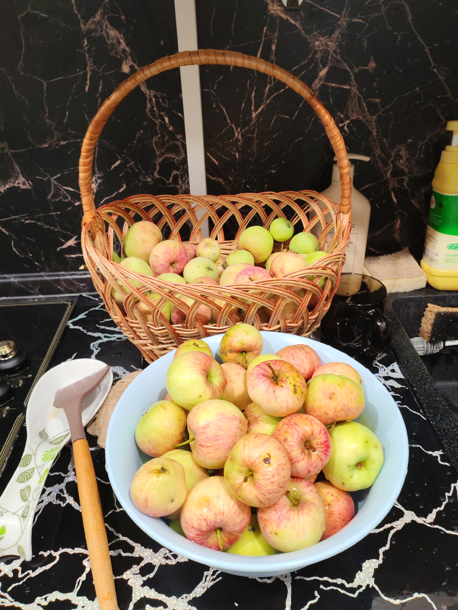 A close-up shot captures a collection of apples displayed in a rustic woven basket and a light blue bowl. The apples, a mix of green and red, appear to be freshly harvested. The background features a black marble-patterned countertop and backsplash, with a white vertical element suggesting a cabinet or appliance. A bottle of yellow dish soap and a sponge are visible to the right, indicating a kitchen setting. To the left, a white spoon rest with a leaf pattern and a wooden-handled utensil add to the domestic ambiance. The scene is lit from above, casting soft shadows on the apples and countertop. The overall impression is one of abundance and home-preparedness, possibly for cooking or preserving. There is no discernible information about the city of Тешевичи, Russia.