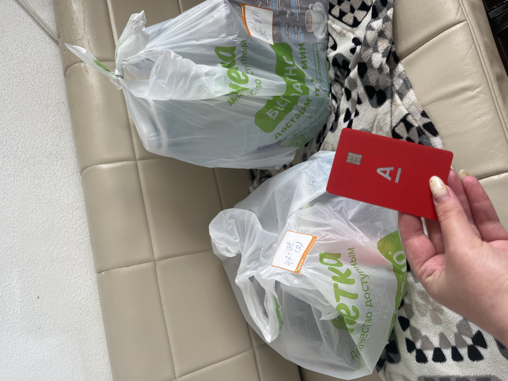 The image shows two plastic grocery bags, one on the left and one on the right, placed on a light-colored, possibly leather, sofa. A hand holding a red card with a white "AI" logo is in the foreground, partially obscuring the left grocery bag and the sofa. The bag on the left has green lettering that reads "МОНЕТКА", which is a Russian grocery store chain, along with "Качество доступным". A handwritten label on this bag says "42-38 (2)". The bag on the right also has green lettering and appears to contain dark items. Both bags are tied at the top. The background consists of a textured, light-colored wall and the sofa. The scene suggests a domestic setting, possibly a living room, in Chelyabinsk, Russia, where the "MONETKA" chain operates.