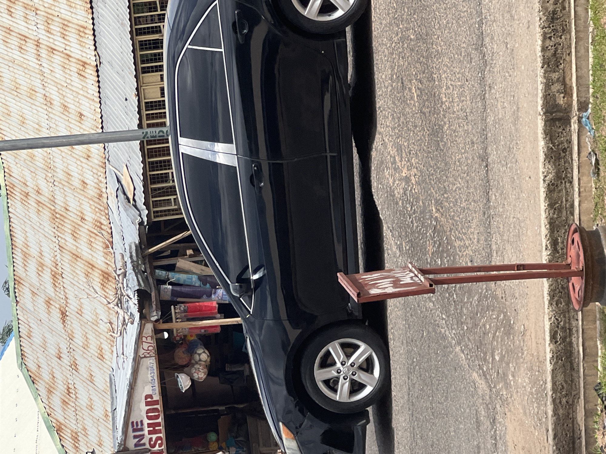 The image shows a black car parked next to a market stall in Jalingo, Nigeria. The market stall has a corrugated iron roof and is filled with various items, including what appear to be fabrics, footballs, and hats. A sign on the stall reads "NE SHOP" and a phone number, "0816543873", is also visible. The car is positioned on a paved road, with a curb and some grass visible to the right. A red metal stand with a white sign is placed near the car. The scene appears to be outdoors during the daytime, with no strong indication of the weather. The overall impression is a snapshot of everyday life in a Nigerian town.