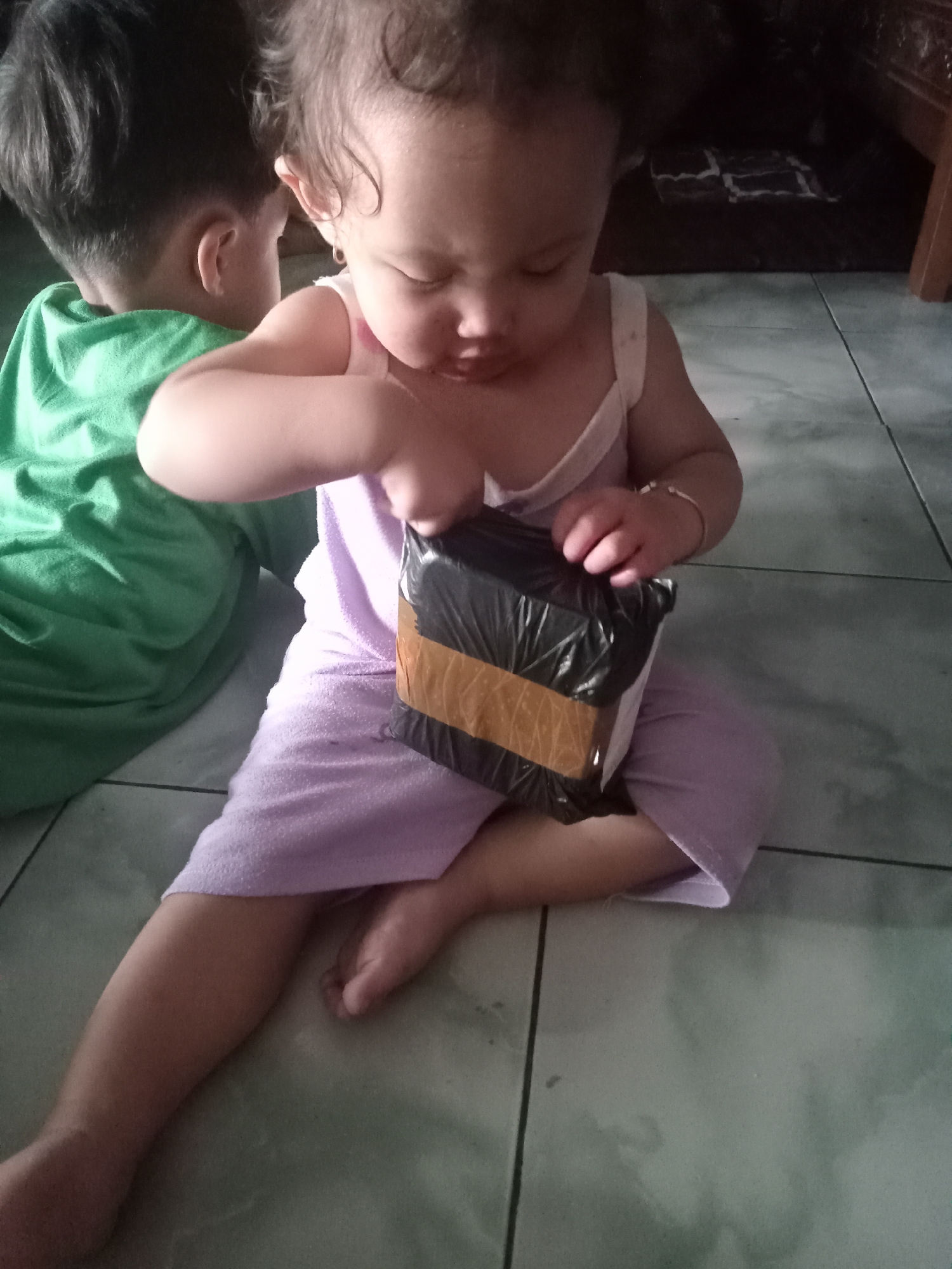 The image depicts two young children indoors, likely in Bungur, Indonesia, with the foreground child intently interacting with a package.
In the center foreground, a baby or toddler, wearing a light purple sleeveless romper, sits directly on a tiled floor. This child has curly dark hair and a small hoop earring visible in their left ear. They are focused on a small, rectangular package wrapped tightly in black plastic and secured with brown packing tape, using both hands to explore or attempt to open it. A thin, silver-colored bracelet is visible on the child's left wrist. The child's expression suggests curiosity and concentration.
To the left and slightly behind, another child, clad in a green t-shirt, sits with their back mostly turned towards the camera, only a portion of their head and shoulder visible.
The setting appears to be an indoor space with light green or grey tiled flooring, featuring distinct dark grout lines. The background on the right shows a dark area, possibly under furniture, with a wooden furniture leg visible at the far right edge of the frame. There is no visible text in the image. The scene conveys a moment of childhood discovery and play.