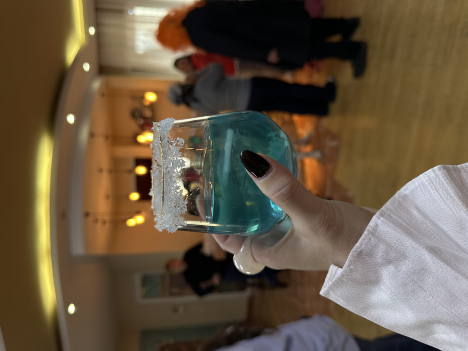 A close-up photograph centers on a left hand, featuring pale skin and manicured, dark, almond-shaped fingernails, holding a clear stemless glass. The individual wears a white, textured sleeve. The glass contains a teal-blue liquid with what appears to be a large ice chunk or multiple ice cubes, and its rim is coated with coarse white crystals.
The background, set in an indoor environment within Tbilisi, Georgia, is out of focus, depicting a social gathering. Several individuals are present: a male figure in a dark long-sleeved top and dark pants stands to the left with hands on hips. To the right, an individual with bright orange, possibly styled or wigged hair, wears a dark garment and is seen from behind. Other blurry figures are partially visible throughout the midground. The room features light wooden flooring and light-colored walls, illuminated by multiple warm-toned pendant lights and recessed ceiling lights. A curved architectural detail is visible on the ceiling, and a sheer white curtain is present on the far right.