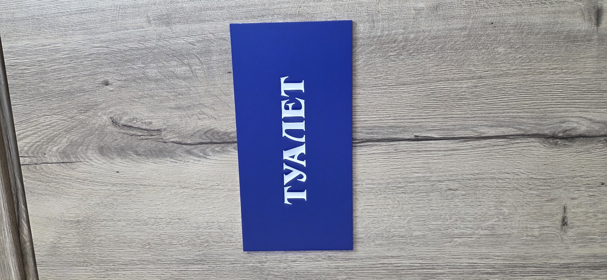 A blue rectangular sign with white text lies horizontally on a light gray wooden surface. The sign is positioned towards the center-right of the frame, occupying about one-third of the image width. The wooden surface displays a prominent grain pattern with varying shades of gray and light brown, suggesting either laminate flooring or a wooden table.
The white text on the blue sign is written in Cyrillic script and reads "ТУАЛЕТ", which translates to "TOILET" or "RESTROOM". The letters are bold and have serifs. The sign appears to be made of a solid material, possibly plastic or painted wood, and is casting a subtle shadow on the wooden background, indicating a light source from above or an angle. No people are visible. The setting is likely indoors, given the appearance of the wooden surface and the sign's nature. The image is well-lit, with no harsh shadows or extreme lighting conditions.