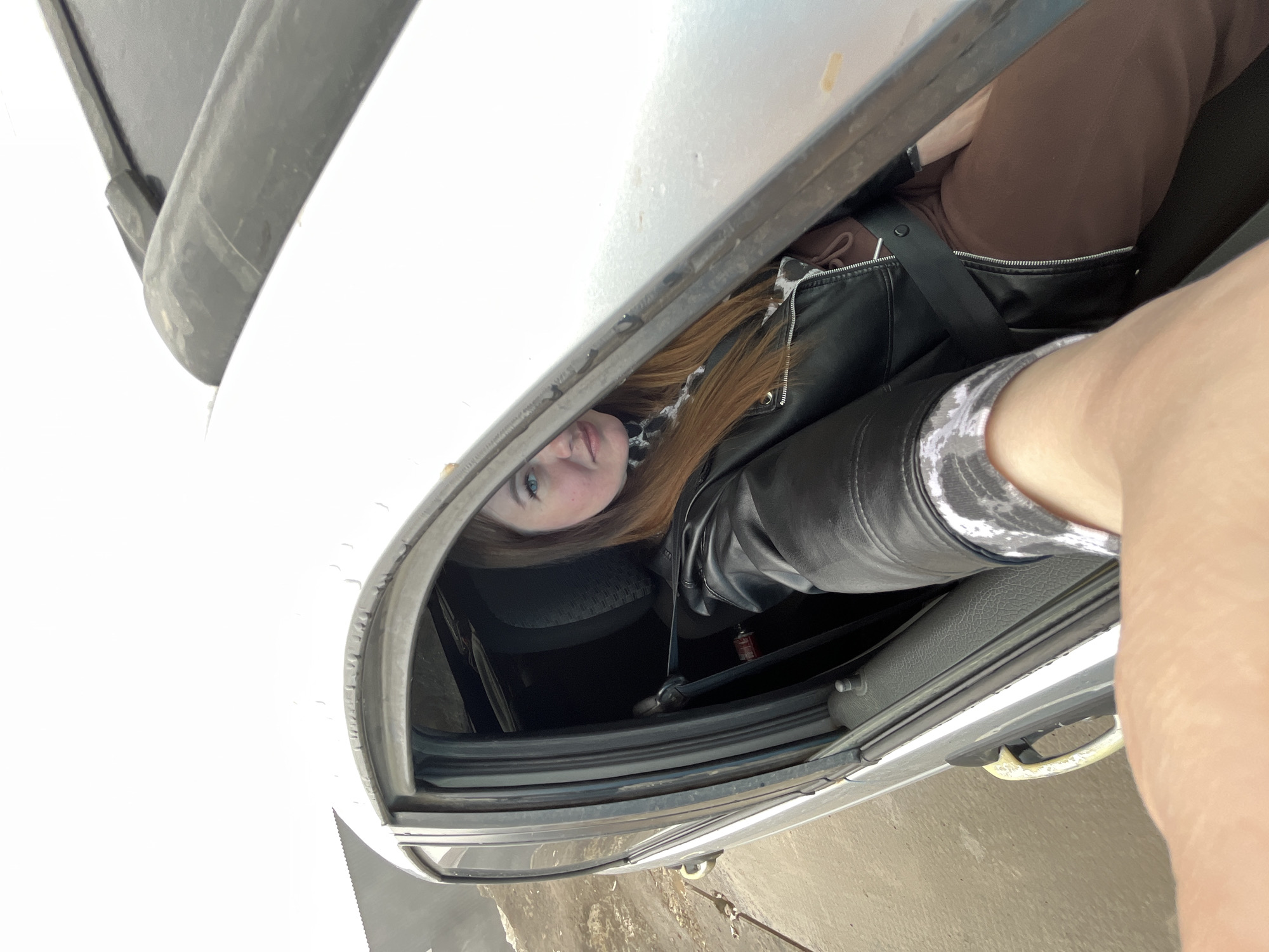 A woman with red hair and blue eyes is inside a white car. She is wearing a black leather jacket and brown pants. The location is the city of Chaikovsky, Russia.