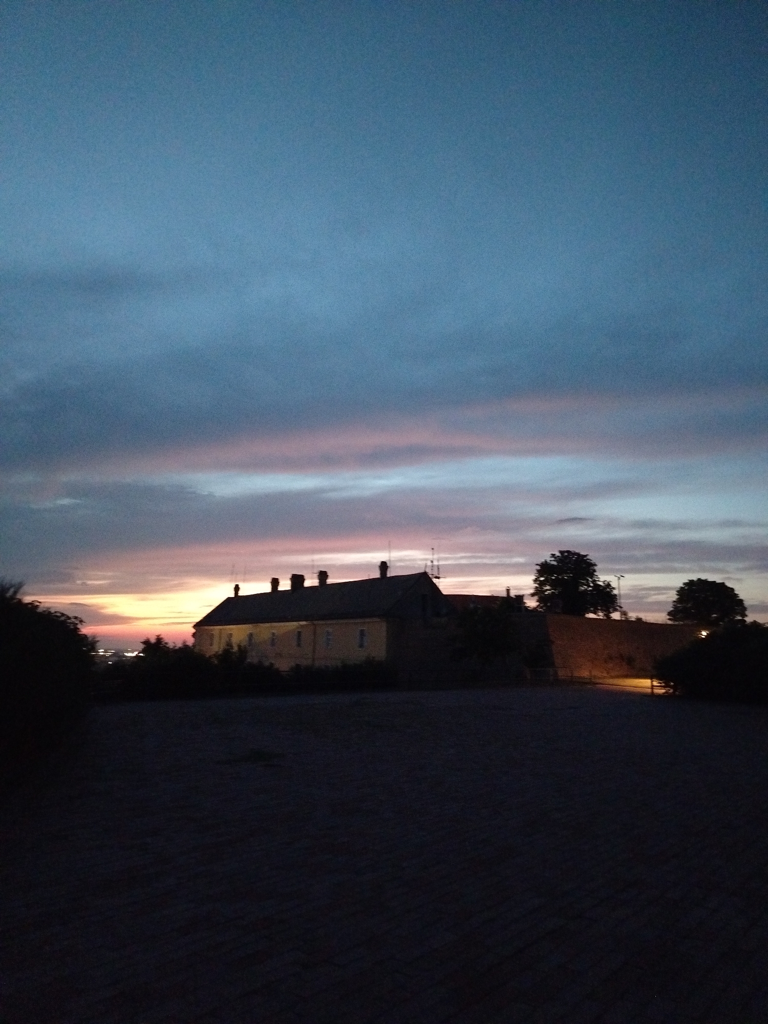 The image captures a twilight scene at the Petrovaradin Fortress in Petrovaradin, Serbia. The sky is a gradient of deep blue at the top, transitioning to softer blues, pinks, and oranges near the horizon, indicating the end of daylight. Silhouetted against the colorful sky is a long, low-lying building with multiple chimneys, likely part of the fortress complex. The building appears to be constructed from stone or brick, with visible windows. To the right of the building, a section of a fortified wall with a warm, illuminated path is visible, suggesting architectural elements of the fortress. Several trees are also silhouetted against the sky, some with dense foliage. In the foreground, a dark, textured surface, possibly a paved area or ground, is visible, with very little detail due to the low light conditions. The overall impression is one of historical grandeur combined with the serene beauty of twilight.