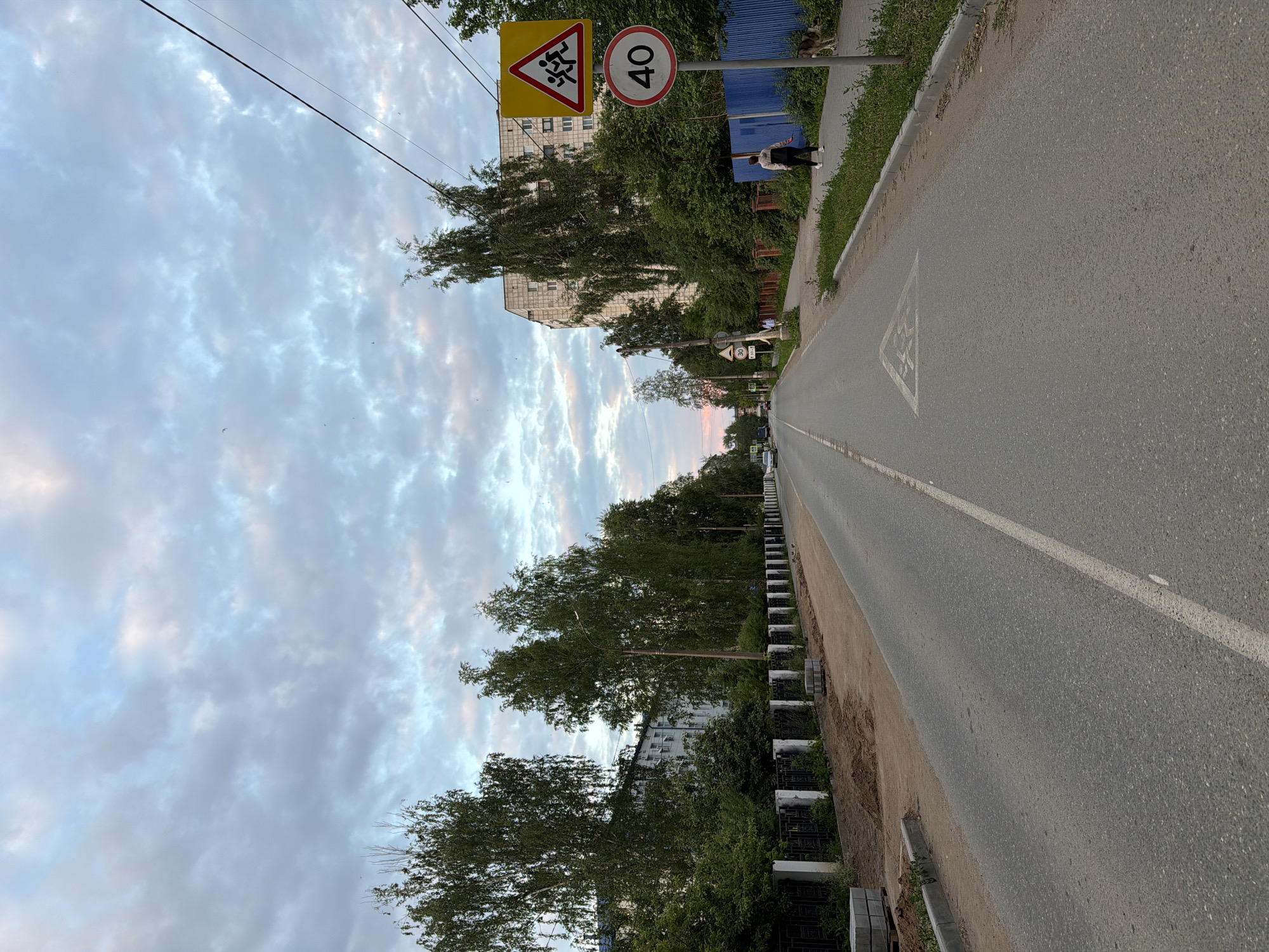 The image depicts a street scene in Chaikovsky, Russia, during dusk. A two-lane asphalt road stretches into the distance, with a white dashed line marking the center. On the right side of the road, a sidewalk runs parallel, bordered by green grass and a blue metal fence. A woman wearing a light-colored tracksuit and white sneakers is walking on the sidewalk, and a dog is nearby. Two traffic signs are visible: a yellow diamond-shaped sign with a depiction of children running, indicating a school zone, and a circular sign with the number "40" in a red border, signifying a speed limit of 40 kilometers per hour. Further down the street, a pedestrian crossing symbol is painted on the road. On the left side of the road, a black metal fence with white posts separates the sidewalk from a grassy area. Beyond the fence, trees line the street in front of apartment buildings. The sky is filled with dramatic clouds, with hues of blue, grey, and pink suggesting the setting sun. Construction materials, including stacks of concrete blocks and loose soil, are visible on the left side of the road, indicating ongoing work. A few cars are present on the road in the distance.