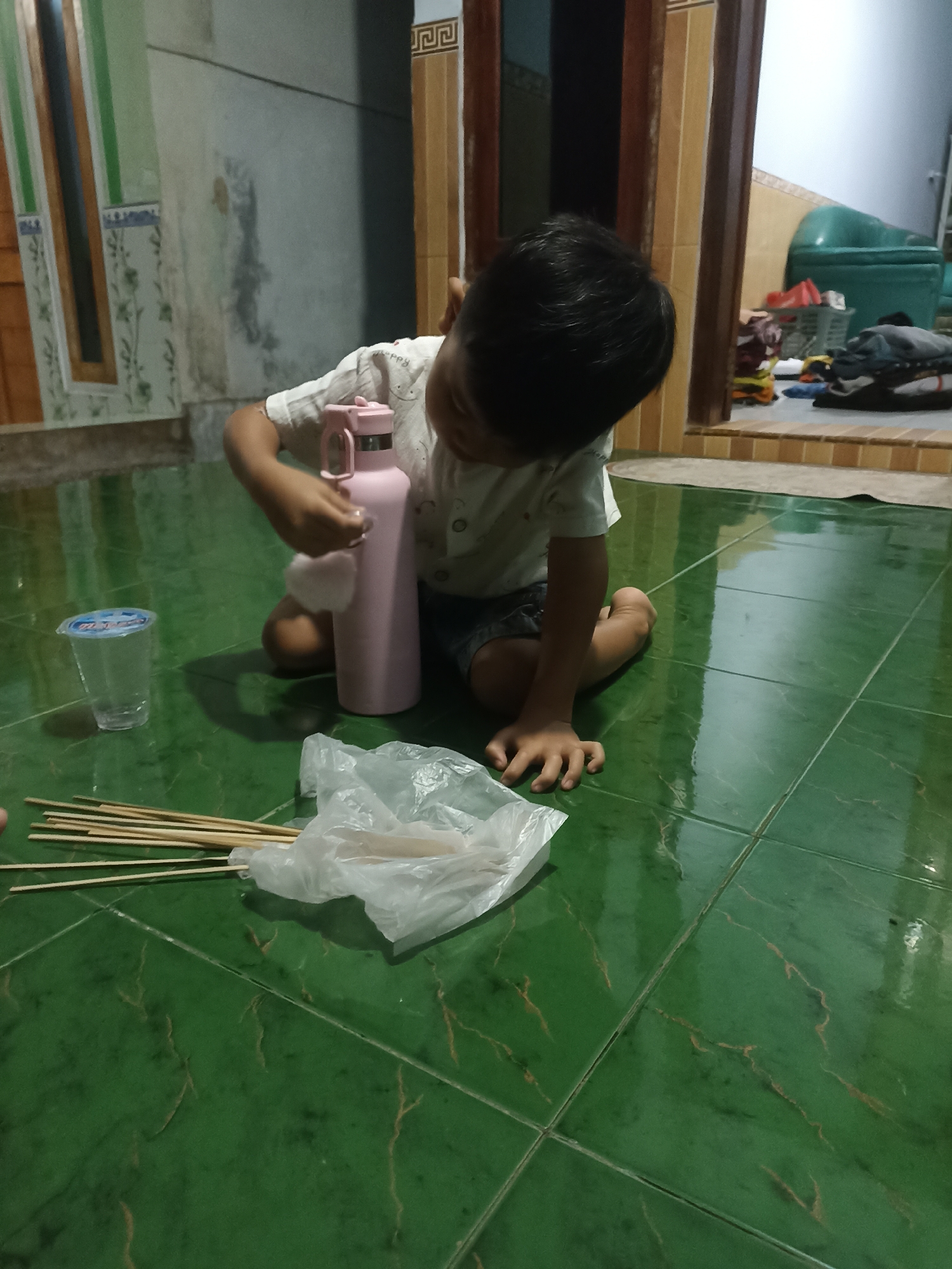 A young child kneels on a green tiled floor in Pandanarum, Indonesia, reaching towards a pink water bottle. The child, wearing a white t-shirt and dark shorts, appears to be interacting with a small white object near the bottle. To the child's left, a clear plastic cup with a blue lid sits on the floor. In front of the child, a bundle of wooden skewers is partially covered by a white plastic bag. The background shows a wall with decorative tiles and a doorway leading to another room where laundry is visible in a basket. The lighting suggests it is indoors, possibly in a home.