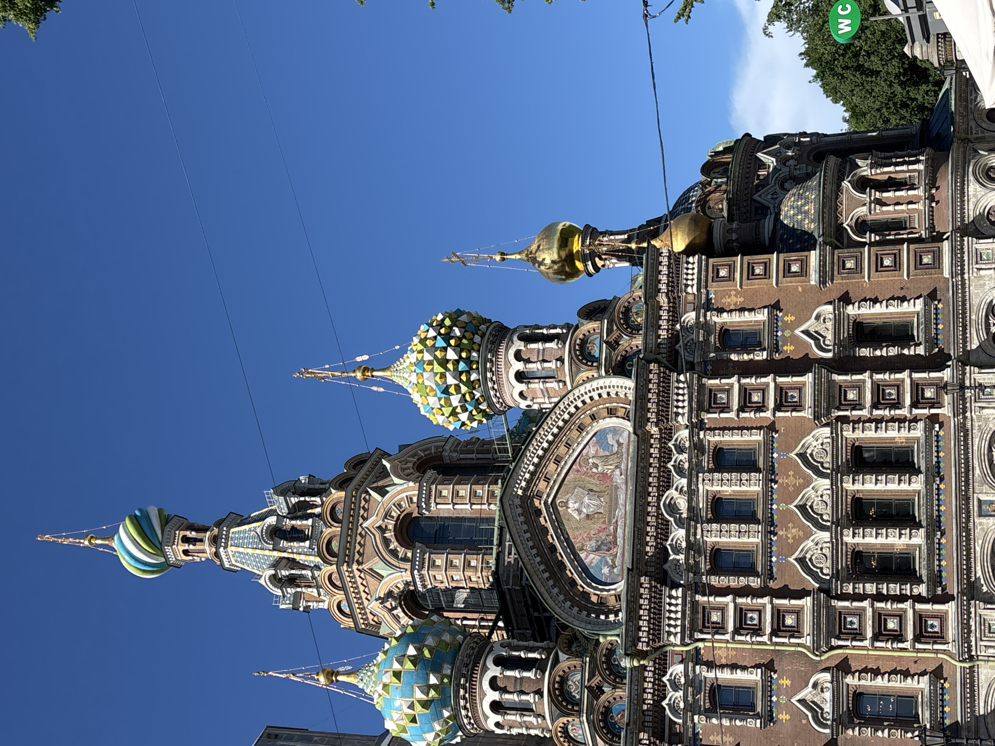 The image displays a close-up, low-angle view of the highly ornate facade of the Church of the Savior on Spilled Blood in Saint Petersburg, Russia, under a clear blue sky. The church's intricate Russian Revival architecture is characterized by its reddish-brown brickwork extensively decorated with white sculpted trim, colored tile patterns, and mosaic detailing.
Multiple domes are visible:
* A central tall dome features a distinctive spiraled pattern of green, white, and light blue, topped with a golden spire and cross.
* To its left, another dome exhibits a geometric pattern of blue, white, and yellow tiles.
* To its right, a similar dome displays a yellow, green, and blue geometric pattern.
* Further right, two smaller golden onion domes with crosses are present, one partially obscured.
A prominent mosaic panel depicting several religious figures, including a central seated figure (likely Christ) flanked by two others, is situated above an arched window on the mid-section of the building. Numerous windows, both rectangular and arched, are visible across the facade, each framed with decorative elements.
Several dark, thin power lines traverse the blue sky in the upper and middle portions of the frame. On the far right, green foliage from trees is visible, partially obscuring the church and a circular green sign with white "WC" letters, indicating a restroom. No people are visible in the immediate foreground.