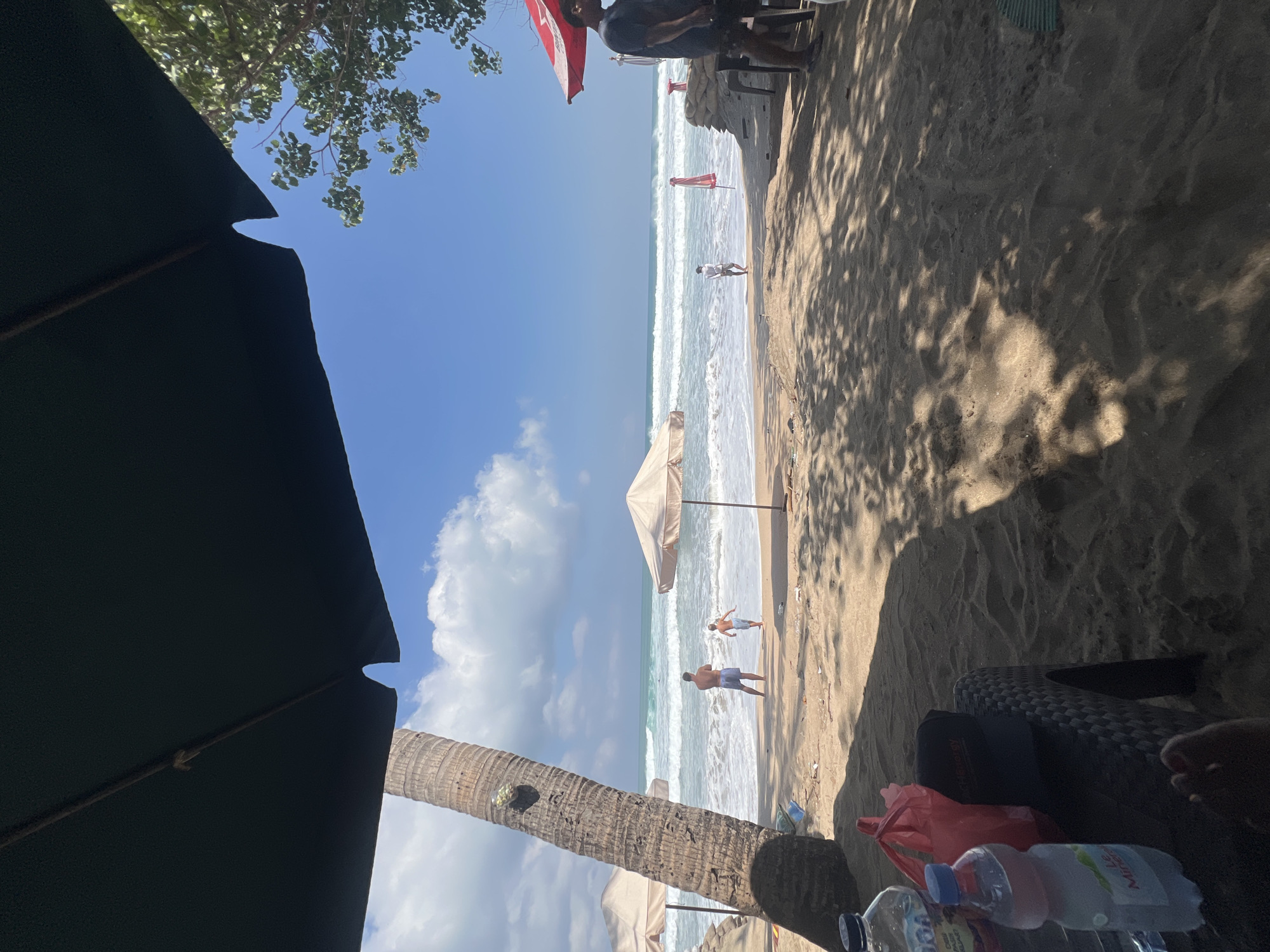 The image depicts a tropical beach scene in Kuta, Indonesia, under a bright blue sky with scattered clouds. In the foreground, a dark umbrella casts a shadow over the sandy beach. To the left, a portion of a palm tree trunk is visible. On a low table in the foreground, there are two plastic water bottles and a red plastic bag.
Further back, several beach umbrellas are set up. Two men and one child are present on the sandy shore near the water's edge. One man stands facing the ocean, and a child runs towards the waves. Another child is also visible near the water. The ocean features moderate waves and a clear horizon.
To the right, another beach umbrella, this one red, is visible. A man stands near a table and chairs on the sand. The sand is a light brown color, and shadows from foliage are cast across it. The overall impression is a relaxed beach environment, typical of Kuta.