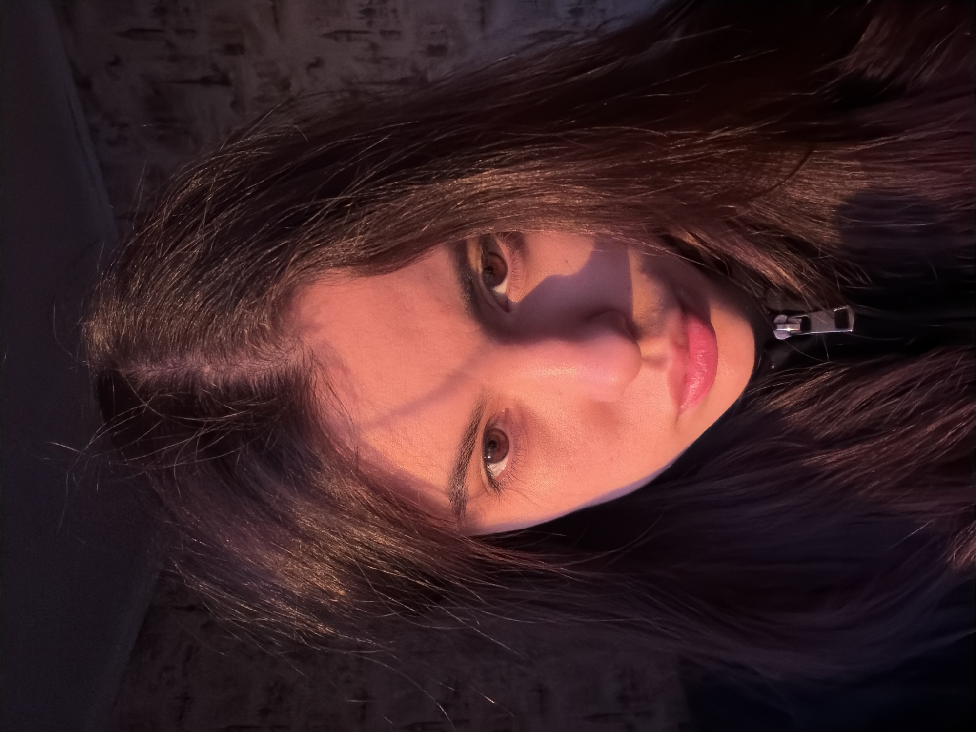 A close-up portrait features a young person, likely female, with long, dark brown hair, captured from an overhead perspective with their head tilted significantly to the right (approximately 90 degrees clockwise from upright). The subject's face is illuminated by warm, directional light, casting prominent shadows on the left side of their face, under their nose, and jawline. Their skin appears fair with warm undertones, highlighted by the golden hour-like glow.
They have deep brown eyes, looking directly at the camera, with well-defined, dark eyebrows. Their nose is delicate, and their full lips have a soft pink tint. Part of a dark, possibly black, zippered garment is visible near their chin and neck, with a metallic zipper pull glinting in the light. Their long, straight to slightly wavy hair is spread around their head, with individual strands catching the warm light, creating a shimmering effect. The background is largely dark and indistinct, suggesting an indoor setting, possibly a wall or soft surface, but lacks specific details.
The overall mood conveyed is intimate and somewhat contemplative due to the direct gaze and lighting. There is no visible text. The photograph appears to capture a posed moment, possibly while the subject is reclining. The location context for this image is Chaikovsky, Russia.