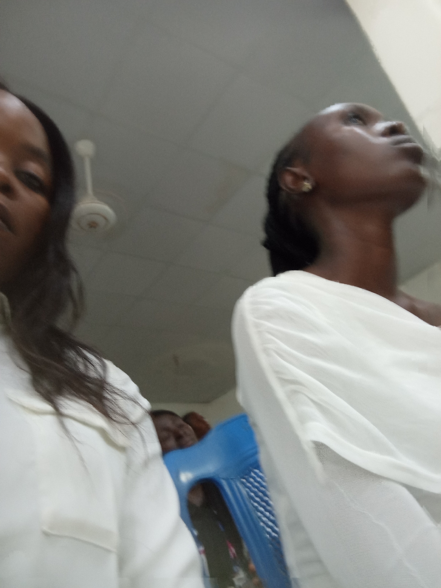 In a dimly lit room, two individuals are captured in a low-angle shot. On the left, a person, partially visible, wears a white top and has dark hair. Their face is mostly out of frame, with only a glimpse of their eye and cheek visible. To the right, another person, facing upwards and to the right, is adorned in a white garment that drapes around their shoulder. They wear a small, gold-toned earring in their visible ear. In the background, a portion of a blue chair is discernible, and a ceiling fan is mounted overhead. The scene appears to be indoors, possibly in Kason, Nigeria.