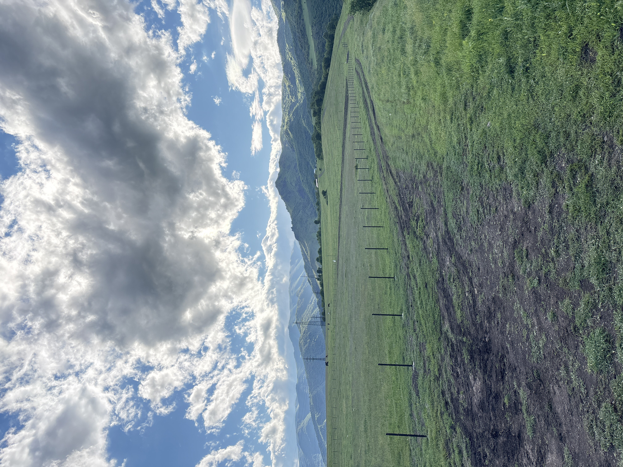 The image shows a landscape with a field to the right and mountains to the left, under a partly cloudy sky. A fence line of wooden posts runs vertically through the image, separating the field. The ground near the bottom appears to be disturbed, showing dirt and less vegetation than the upper portions of the field. In the distance, power lines can be seen. The location is in a mountainous area of Alaverdi, Armenia. There are no visible people in the image.