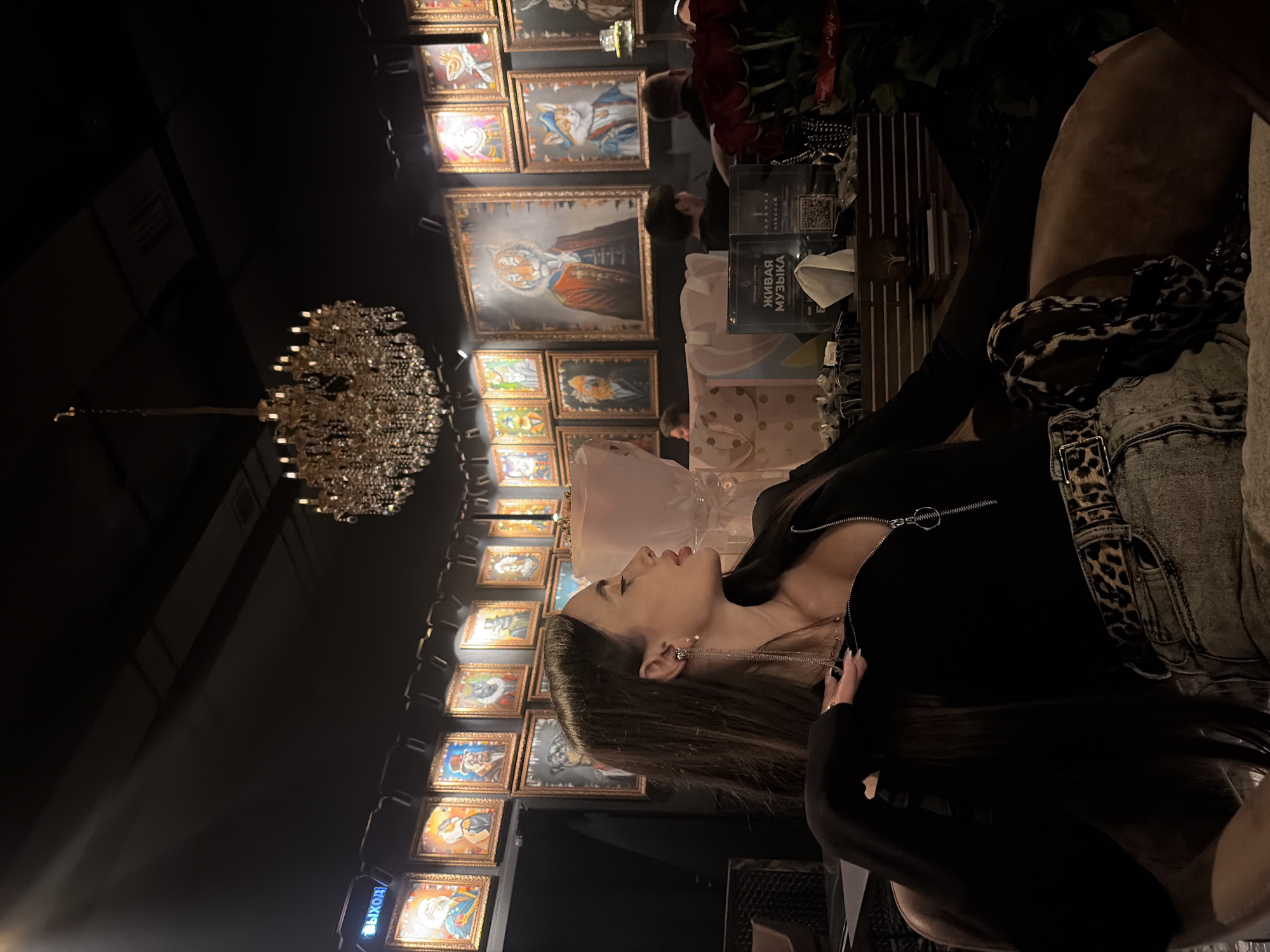 The image shows a young woman indoors, seated at a table with a low-cut black top and a denim jacket with a leopard print belt, in what appears to be an upscale restaurant. Her head is turned to the side, facing a wall adorned with framed paintings. Above her, a large chandelier hangs from the ceiling. In the background, other patrons are visible. The environment suggests a lavish interior. The location is likely Saint Petersburg, Russia, judging from the Cyrillic script on signage in the background.