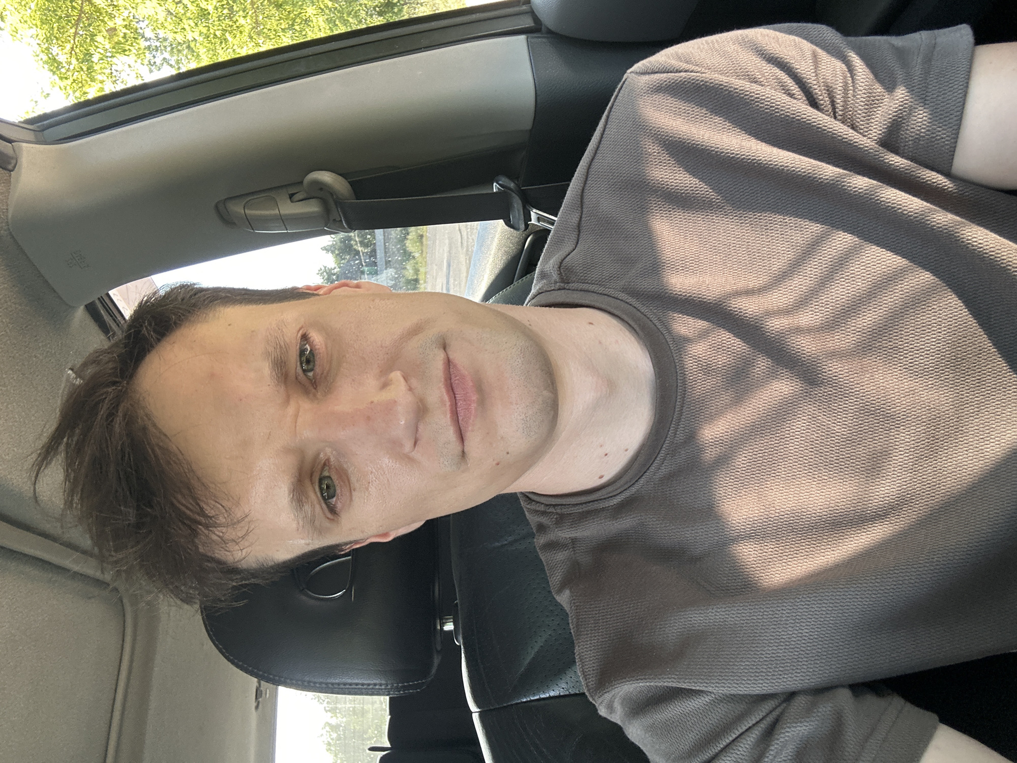 The image is a selfie of a man sitting in the driver's seat of a car. He has light skin, short brown hair, and green eyes. He is wearing a gray t-shirt. The interior of the car is visible, including the headrest of the passenger seat and the seatbelt mechanism. The exterior of the car shows trees and a road. The lighting suggests it is daytime, possibly late afternoon due to the shadows. The man's expression is neutral. There are no other people or animals visible in the image. The location is identified as Makiv, Ukraine.