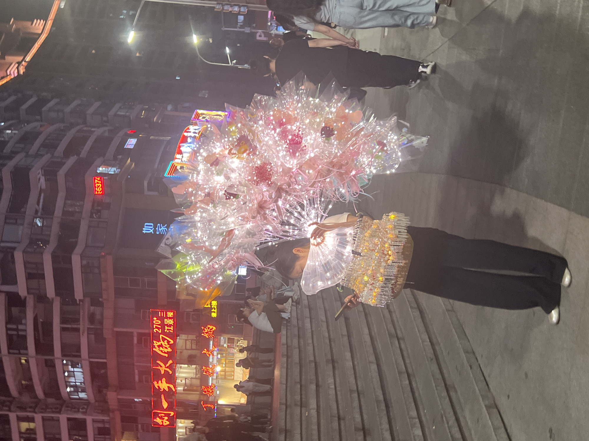 A street vendor stands on steps in the city of Yuzhong District, China, holding an assortment of illuminated balloons and a small fan. Buildings with neon signs are visible in the background. Pedestrians walk by.