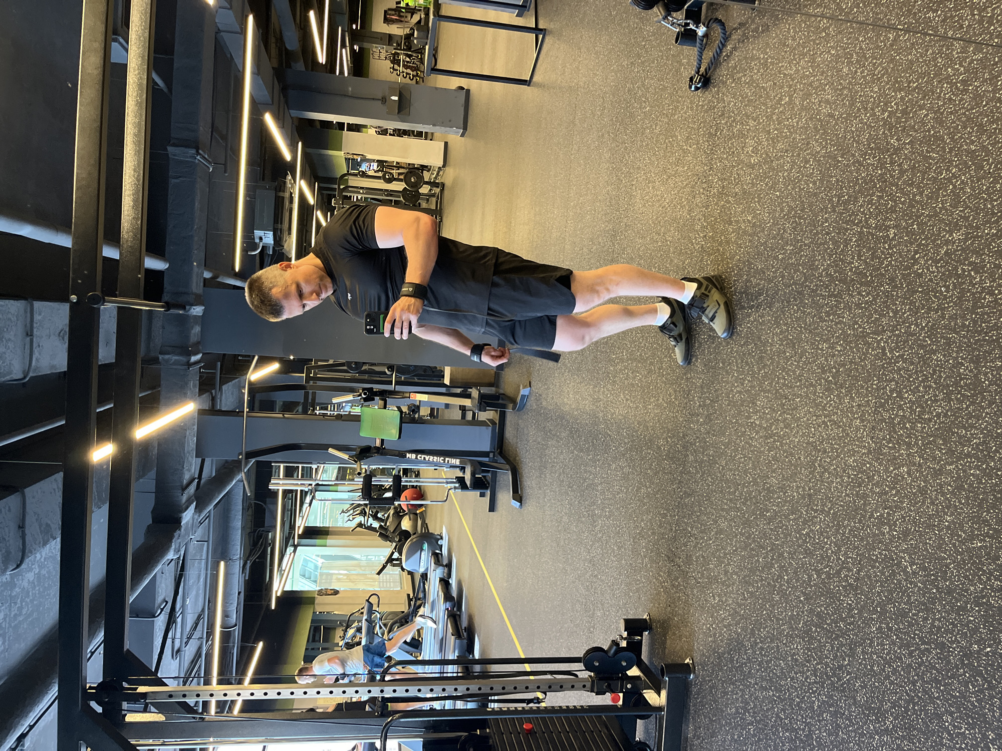 The image depicts an indoor gym facility in Sochi, Russia. A man, approximately in his 40s, with short, greying hair, is positioned in the foreground-right, taking a selfie with a black smartphone held in his right hand. He is wearing a black short-sleeved athletic shirt with a small white logo on the left chest, black athletic shorts, white ankle socks, and dark grey/olive athletic shoes with black strap closures. Black wrist wraps are visible on both wrists. He is looking down at his phone, with his body slightly angled to the left.
The gym features dark grey rubberized flooring flecked with white, with a distinct yellow line running horizontally across the mid-ground. In the background-left, another man is partially visible, wearing a light-colored t-shirt and blue shorts, running on a treadmill. Several other treadmills and various cardio and strength training machines are visible throughout the background, including ellipticals and weight stations, some with green upholstered pads.
The ceiling is exposed, revealing dark grey ventilation ducts, pipes, and multiple linear LED light fixtures that emit a warm white glow. The walls are predominantly dark grey, with some sections appearing reflective or containing large windows. On the far right, a black-framed table-like structure is present. A cable machine with weight stacks is partially visible on the far left of the frame. The overall environment is well-lit, spacious, and appears to be a modern fitness center.