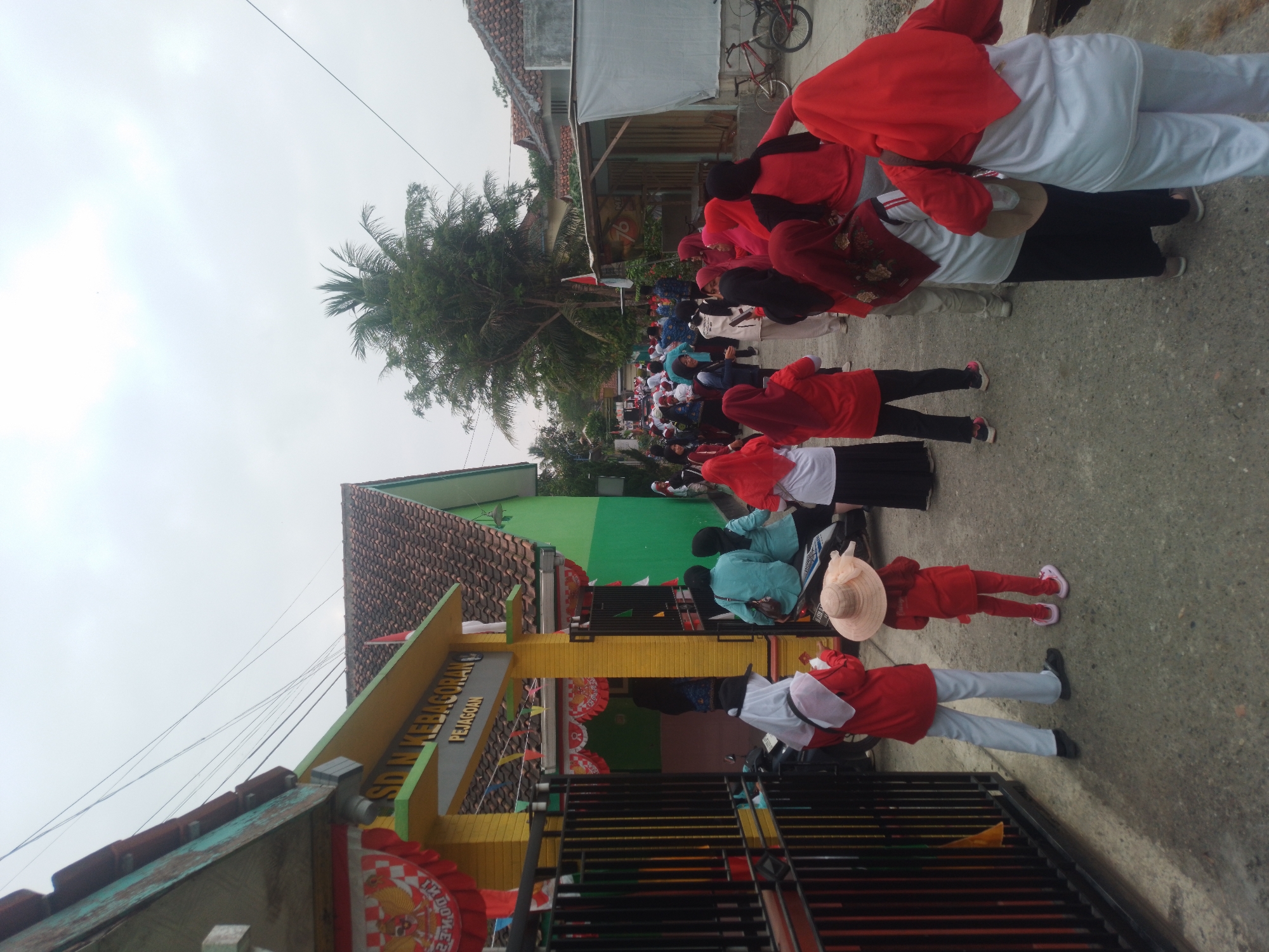 The image shows a street scene in Kebagoran, Indonesia, in front of a school building. The school entrance has a yellow archway with "SD N KEBAGORAN PEJAGOAN" written on it. Several people, mostly women and children, are gathered on the street, many wearing red attire. Some are facing the school, while others are walking away. There is a fence on the left side of the image and a green building to the right of the school entrance. A coconut tree is visible in the background, along with other buildings and a cloudy sky. A bicycle is parked near a structure on the right side of the image. The ground is paved with asphalt.