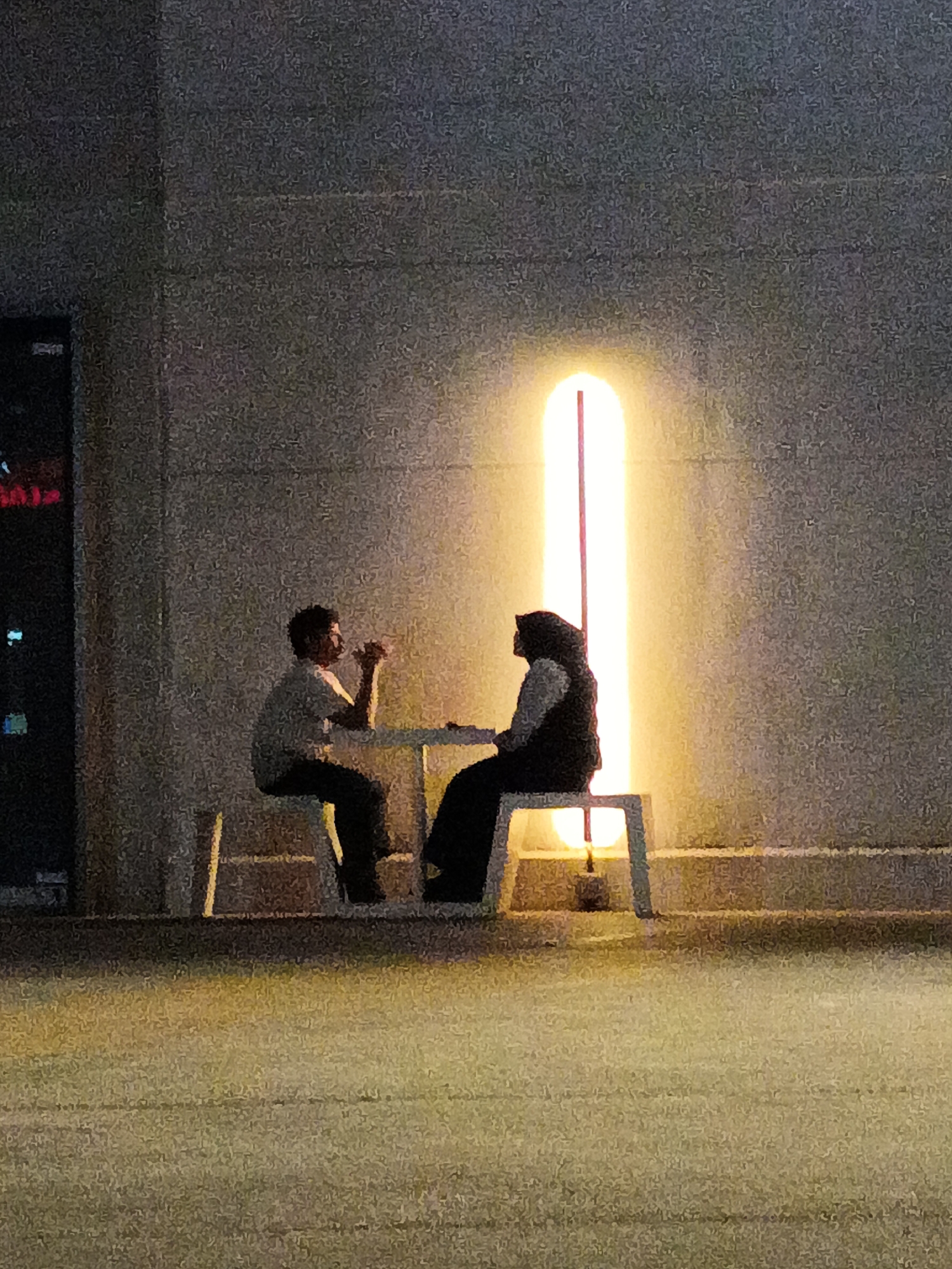 In Kuala Lumpur, Malaysia, two individuals are seated at a white table at night, positioned in front of a textured concrete wall illuminated by a tall, vertical light fixture emitting a warm yellow glow. The person on the left, a male wearing a light-colored t-shirt and dark pants, is gesturing with his hands. Across from him, the person on the right, a female wearing a hijab and a dark vest over a lighter garment, is also interacting, her hands visible on the table. The setting appears to be an outdoor area, possibly a cafe or public space, with a dimly lit asphalt-like surface in the foreground reflecting the ambient light.