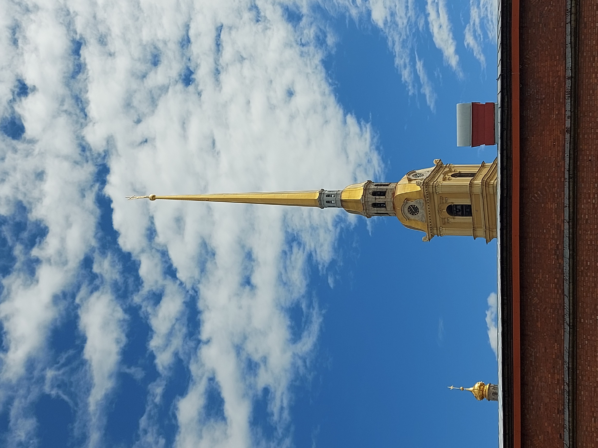 The image shows a view of the Peter and Paul Cathedral in Saint Petersburg, Russia, from a ground-level perspective. The gold spire of the cathedral dominates the frame against a bright blue sky with scattered white clouds. A portion of a brick building is visible on the right edge, suggesting the photograph was taken from within the vicinity of the Peter and Paul Fortress. The lower golden spire of another nearby building is also visible.