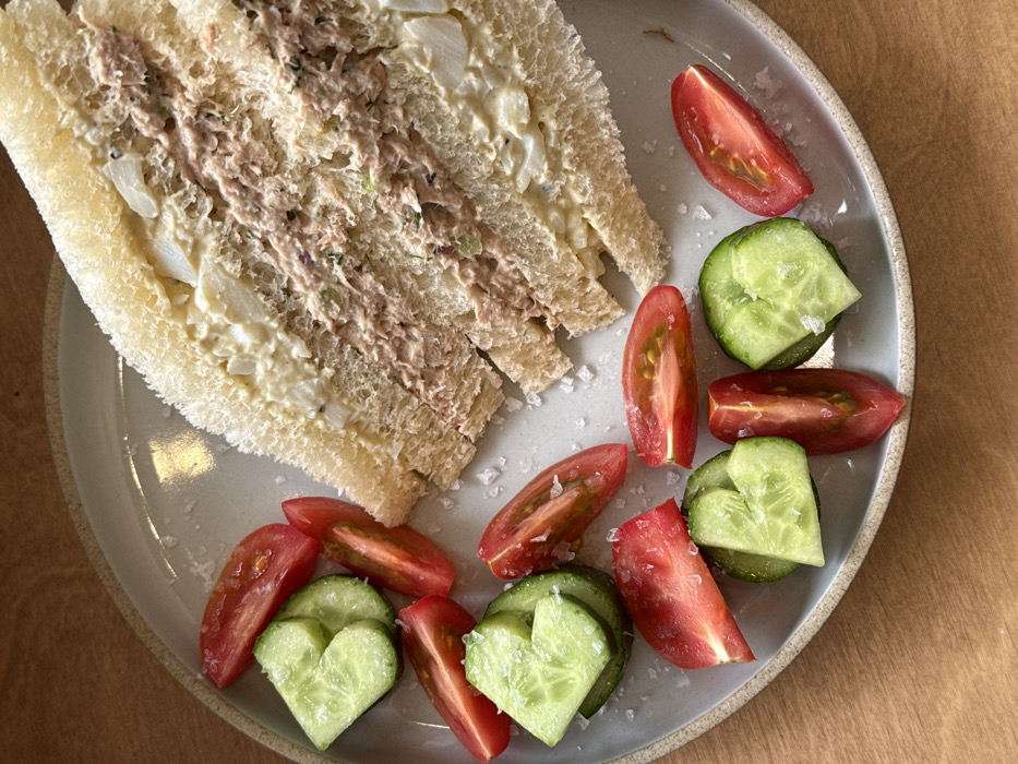 The image displays an overhead view of a meal presented on a light gray ceramic plate with a textured, unglazed rim. On the right side of the plate, two triangular halves of a sandwich are arranged. The sandwich is made with white bread, and its crusts have been removed. The filling comprises two layers: an upper layer of chunky egg salad and a lower layer of tuna salad mixed with mayonnaise, celery, and possibly dill. To the left and bottom of the plate, approximately ten slices of green cucumber and eight red tomato wedges are scattered. Several cucumber slices are cut into heart shapes. Coarse white salt crystals are visible on the vegetables and the plate surface. The plate rests on a light brown wooden surface. No individuals are present, and no specific environmental details are visible to determine a precise location within Tel Aviv-Yafo, Israel.