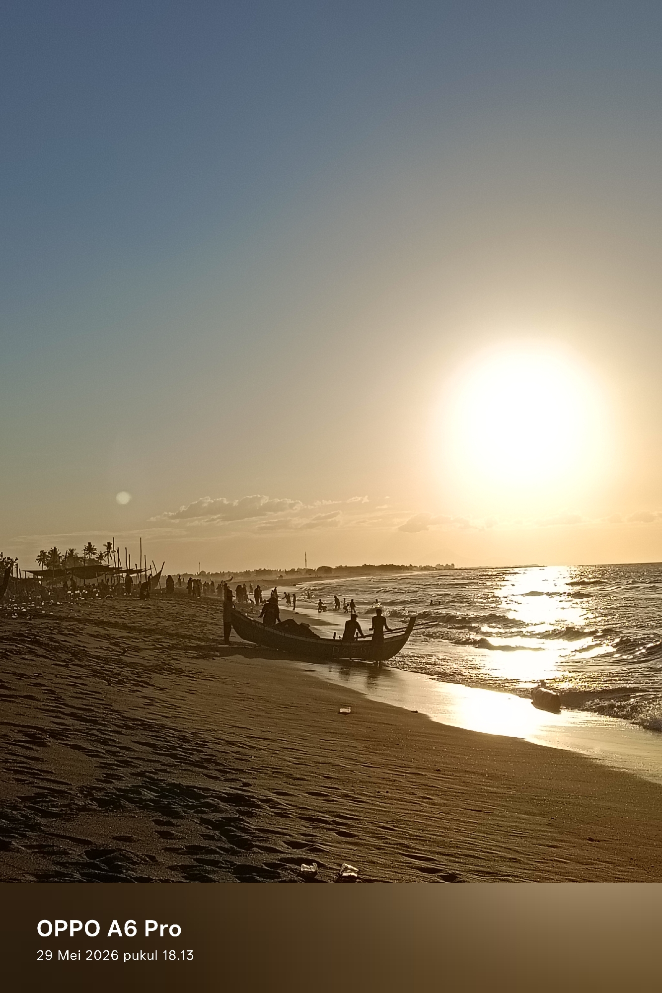 The photograph shows a beach scene at sunset in Rheumbaroh, Indonesia. The sky is clear blue, transitioning to orange near the horizon where a bright sun is positioned. The sun's reflection creates a shimmering effect on the ocean surface. A traditional wooden boat is positioned near the shoreline, with several people standing inside and around it. Silhouetted figures are visible along the beach and in the water. In the background, there are trees and what appear to be simple structures. The sand is textured with footprints and other markings. Some trash is visible on the beach. In the lower portion of the photo, text indicates the image was captured on an OPPO A6 Pro on May 29, 2026, at 18:13.