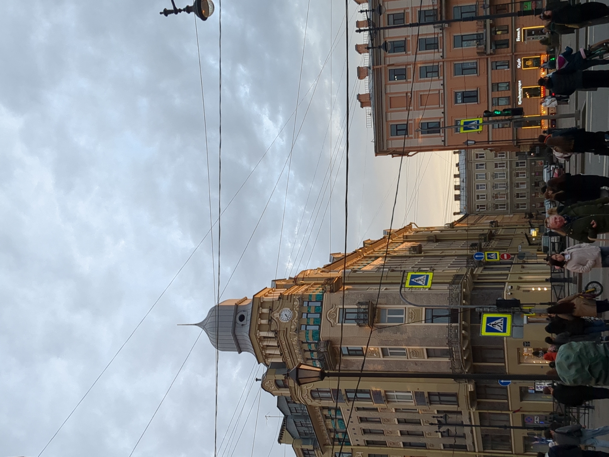 The image captures a street scene in Saint Petersburg, Russia. The architectural style of the buildings suggests a historic urban setting. There is a building with a distinctive tower and clock on its facade. Several overhead electrical wires crisscross the sky. Pedestrians are present, crossing the street at a designated crosswalk. Traffic signs are visible, indicating pedestrian crossings. The sky is overcast.