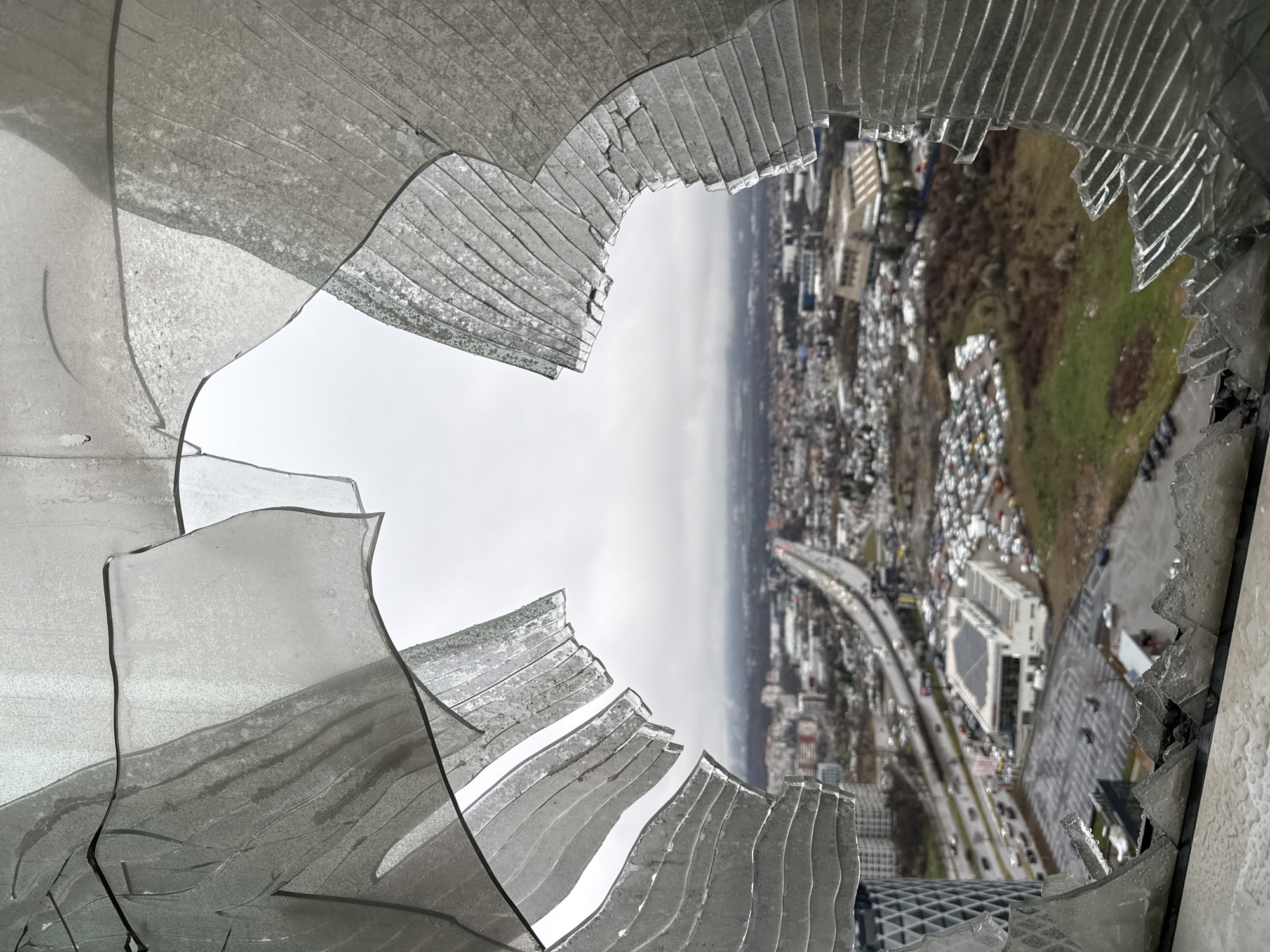 The image presents an elevated, wide-angle view of a city through a severely damaged windowpane. The foreground is dominated by large, jagged fragments of shattered glass, which are bent and fractured, particularly along the top, left, and right edges of the frame. These fragments show multiple layers of breakage and some degree of delamination, suggesting safety glass or similar material, with water droplets visible on their surfaces.
Through the irregular opening created by the broken glass, an urban landscape identified as Sofia, Bulgaria, extends to the horizon under an overcast, grey sky. A prominent multi-lane highway or major thoroughfare runs diagonally through the middle ground, extending into the distance from the upper right to the lower left. Vehicle traffic is visible on this road, with numerous cars appearing as streaks of light.
To the left of the highway, a modern, high-rise building with a distinctive geometric or lattice-patterned facade is visible. Further into the urban sprawl, numerous other buildings of various sizes and architectural styles populate the scene. Several large parking lots, one containing a dense collection of vehicles next to a commercial building with solar panels on its roof, are clearly discernible. Patches of brown and green vegetation are interspersed throughout the urban development. No individuals are visible in the scene. The overall environment suggests an elevated vantage point on a cloudy day, overlooking a bustling city area.