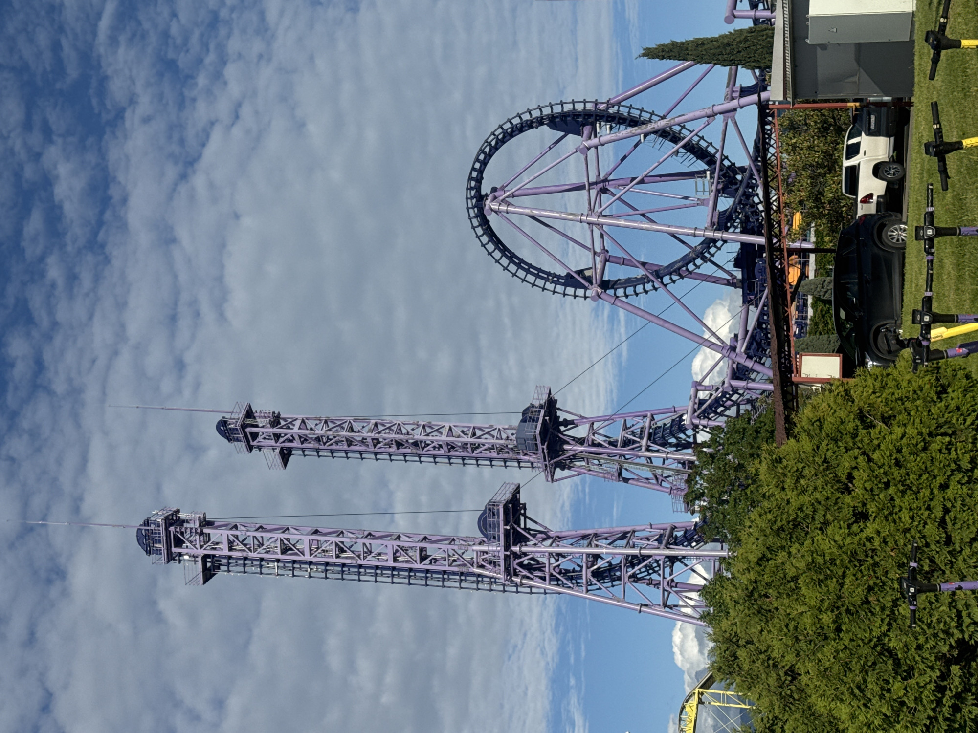 The image shows a purple and black roller coaster with a large circular loop. There are also two tall vertical lift structures. The coaster is located at the Sochi Park in Sirius, Russia. A white pickup truck and a black car are parked near the coaster. There are also scooters nearby. The sky is blue with some clouds. A green bush is in the foreground.