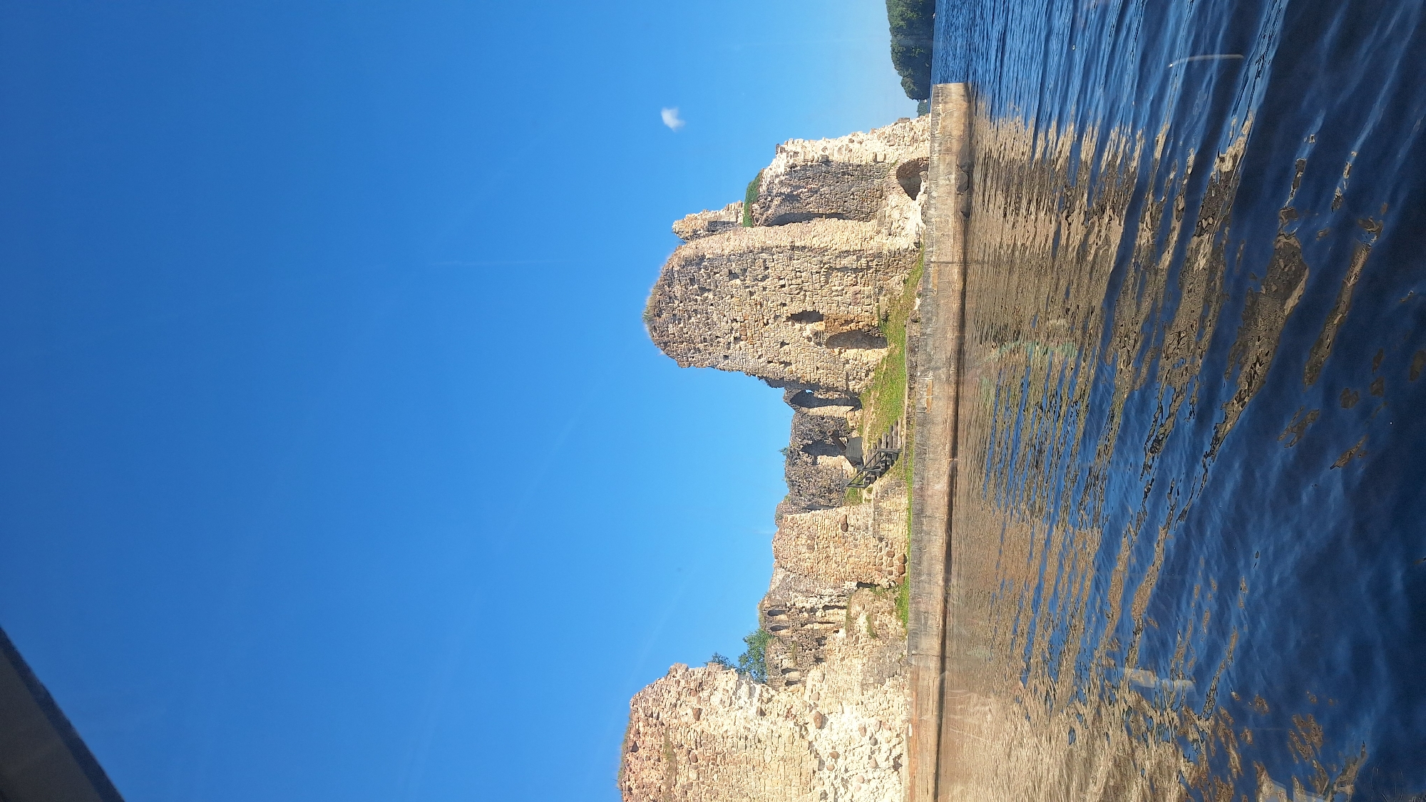 The image depicts the ruins of Koknese Castle, situated on the Daugava River in Koknese, Latvia. The ancient stone structure, characterized by its weathered walls and surviving sections of towers, stands prominently on a small island or peninsula. The castle ruins are bathed in sunlight, casting shadows on the textured stone. Surrounding the ruins, there is a concrete embankment, and beyond that, the calm, blue waters of the Daugava River reflect the sky and the castle. Lush green grass grows on the upper levels of the ruins, indicating the passage of time and the integration of nature with the historical site. The sky above is a clear, vibrant blue with a few wispy clouds. The overall scene evokes a sense of history and natural beauty, with the imposing ruins serving as a stark reminder of the past against the backdrop of a serene riverside landscape.