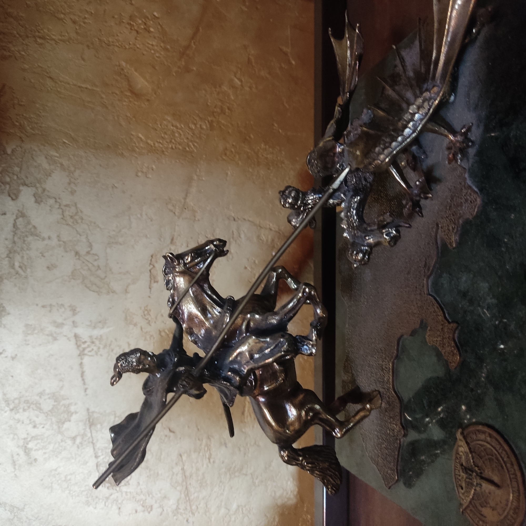 This media file depicts a bronze sculpture of a knight on horseback engaged in combat with a dragon. The knight is in mid-action, with his lance aimed at the dragon. The horse is rearing up, its front legs in the air, as if startled by the dragon. The dragon is also depicted in a dynamic pose, with its wings spread and its mouth open, as if roaring. The sculpture is highly detailed, with intricate rendering of the musculature of the horse and dragon, as well as the armor and weaponry of the knight. The background appears to be a textured wall, possibly stone or plaster, with a subtle color variation suggesting it might be illuminated by artificial light. The overall impression is one of epic struggle and heroic valor. The scene is set indoors, with the lighting suggesting it could be day or night. The lack of any discernible text in the image makes it difficult to ascertain any specific location or time. The metallic sheen of the bronze suggests a polished finish, indicating the sculpture is likely a decorative piece.