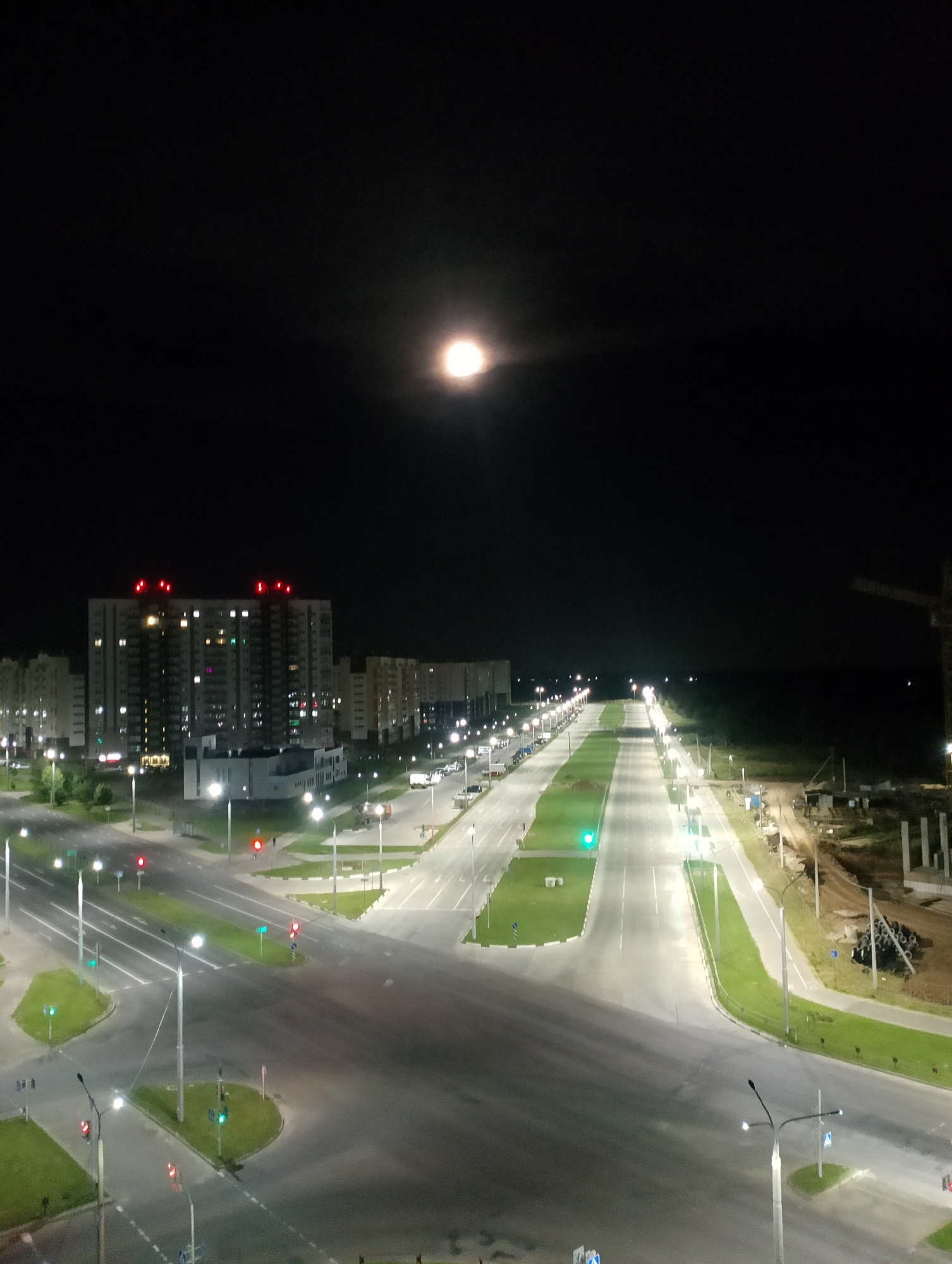 The image is a nighttime aerial view of a newly developed urban area. A wide, well-lit street stretches into the distance, flanked by green medians and modern streetlights. On the left, apartment buildings are illuminated, some with red warning lights on their rooftops. On the right, a construction site is visible with a crane and building materials. The sky is dark, with a bright moon and halo prominently displayed. The scene suggests a planned residential development, possibly in the early stages of occupation, with ongoing construction. The location is identified as Vitsebsk, Belarus. There are no individuals or specific actions depicted, only the static environment.