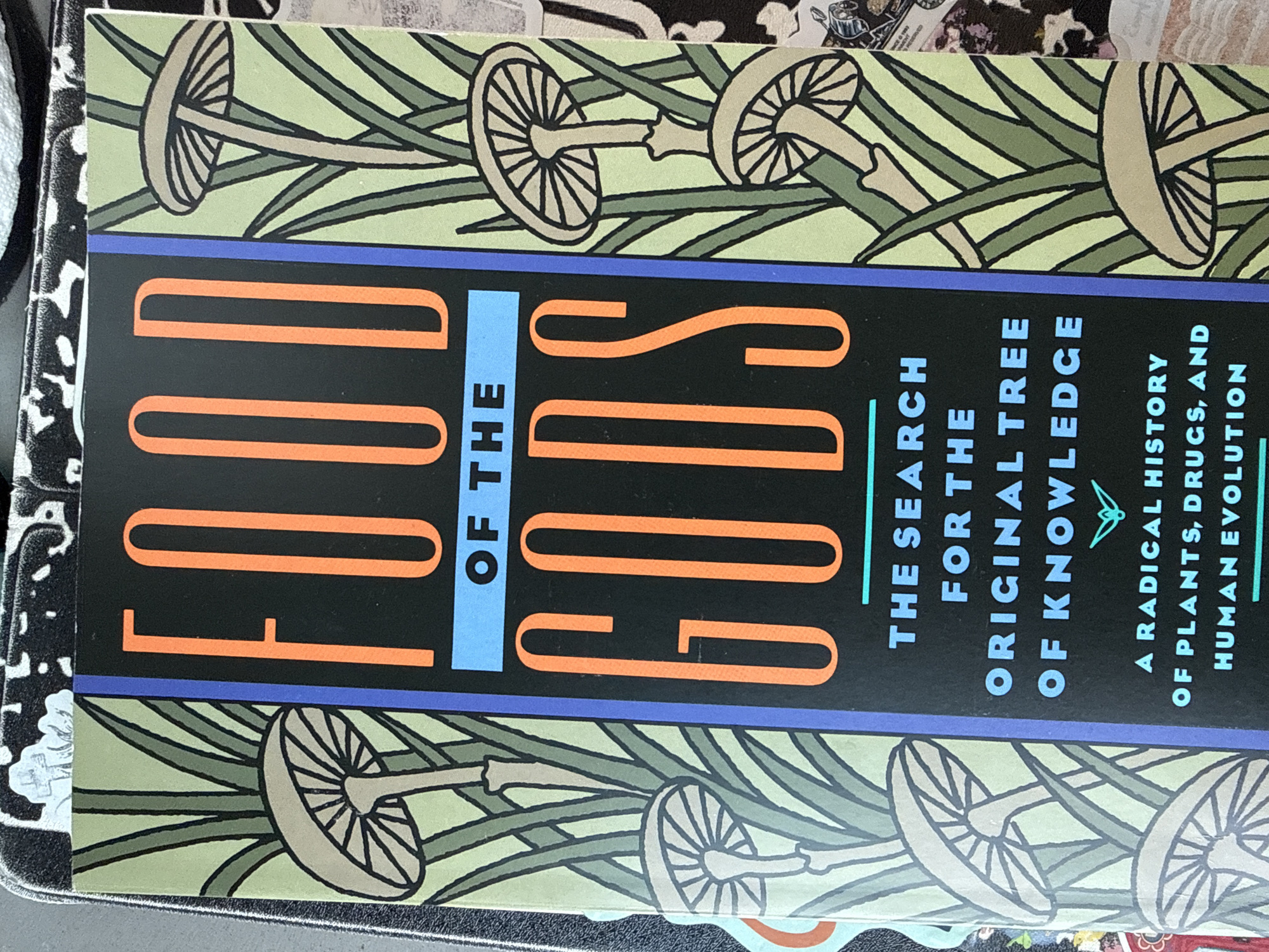 Here is a description of the image:
The image shows a close-up of the book "Food of the Gods" by Terence McKenna. The cover features illustrations of mushrooms and plants on a light green background, with a dark blue border around the title. The text "FOOD OF THE" is in large orange letters, with "OF THE" in light blue. Below the title, it reads "THE SEARCH FOR THE ORIGINAL TREE OF KNOWLEDGE" in a light blue font. There's additional text stating "A RADICAL HISTORY OF PLANTS, DRUGS, AND HUMAN EVOLUTION" in white font.