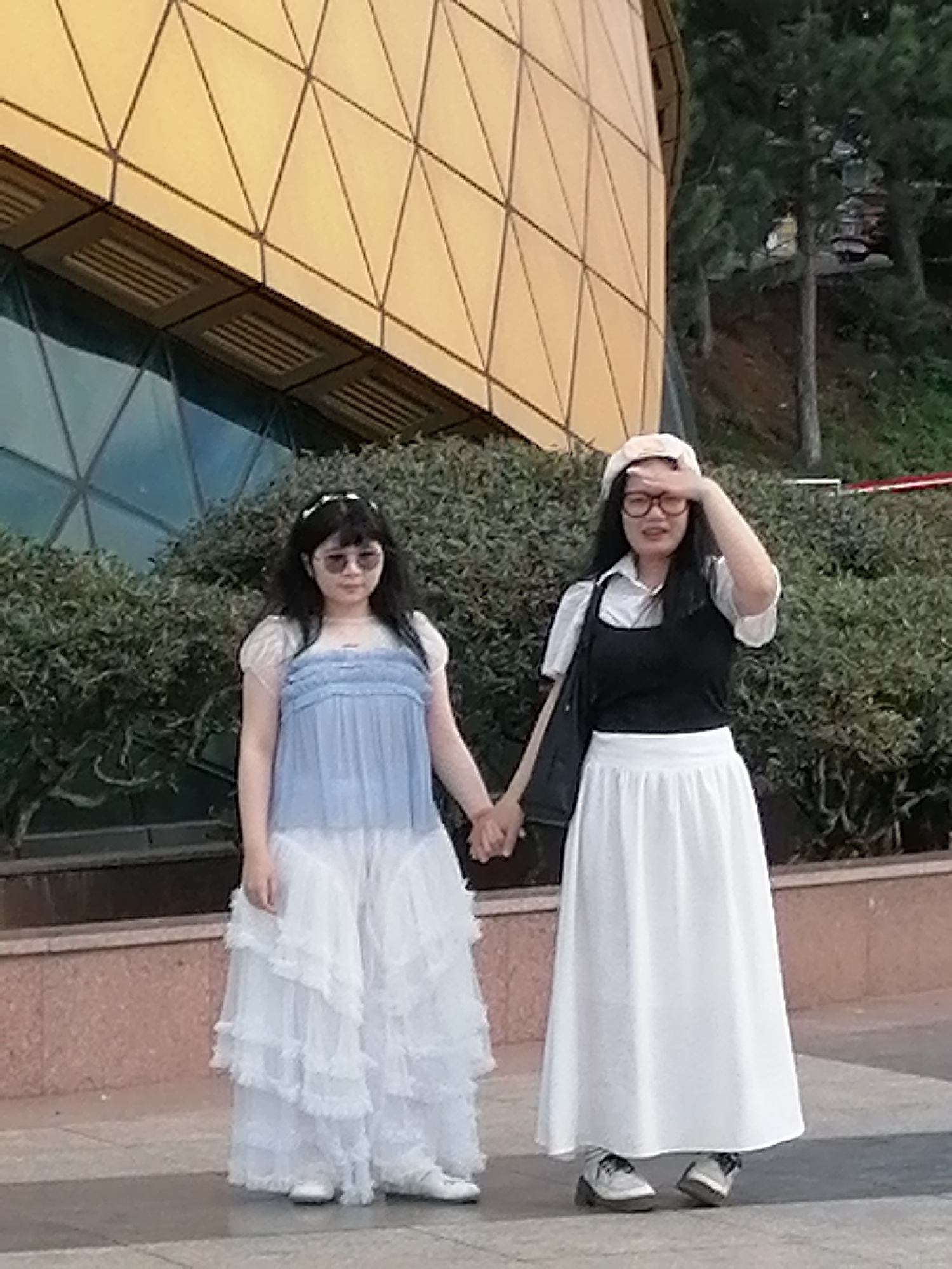 Two young women are standing outdoors, holding hands. The woman on the left wears a light blue, sheer top with ruffled sleeves and a white, layered skirt. She has dark hair and is wearing sunglasses and a headband. The woman on the right wears a white collared shirt, a black sleeveless vest, and a long white skirt. She has dark hair, glasses, and is wearing a light pink beret. She is shielding her eyes with her hand. They are standing in front of a large, modern building with a golden, geometric facade and a large glass window. Behind the building, there are trees and a hint of foliage. The ground is paved. This scene appears to be in front of the Da Lat Cable Car Station in Da Lat, Vietnam, given the distinctive architecture of the building.