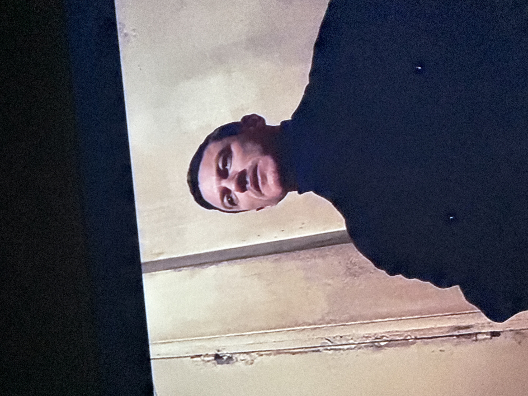 The image shows a man from a low-angle perspective, suggesting he is looking down at the viewer. He has short dark hair and is wearing a dark turtleneck sweater or shirt. The background is a beige wall with horizontal trim. The left side of the image shows part of a television. There is no indication of Mytishchi, Russia, or any other specific location.