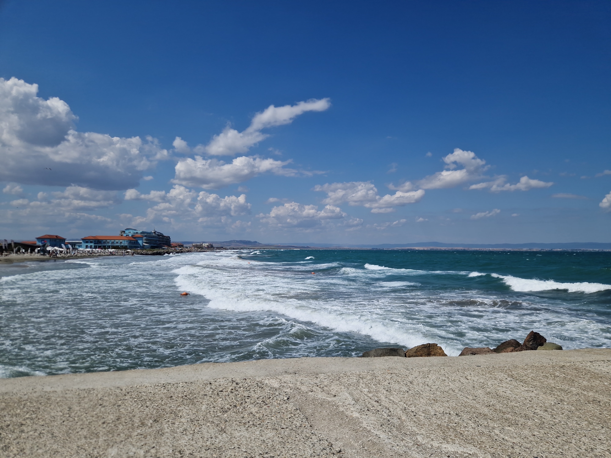 The image depicts a coastal scene in Pomorie, Bulgaria, facing the Black Sea. In the foreground, a textured concrete embankment occupies the lower portion of the frame. Beyond this, the turbulent, greenish-blue waters of the sea churn with white-capped waves. An orange buoy is visible in the water, indicating a swimming area or marker. Along the coastline in the middle ground, a resort complex with distinctive turquoise and orange architecture is situated, backed by a beach with scattered umbrellas. The horizon line is marked by a low, hazy landmass in the distance. The sky above is a vibrant blue, populated by scattered white cumulus clouds. A single bird is visible in flight against the sky on the left side of the image. The overall impression is of a bright, active day at the seaside.