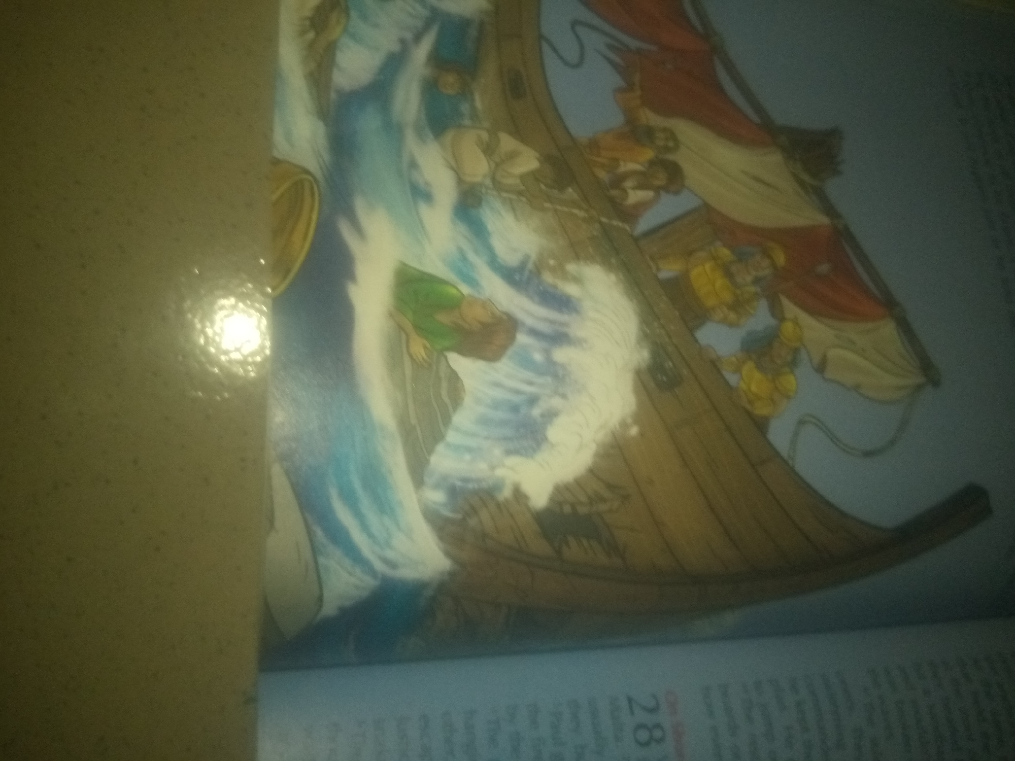 The image shows a close-up, rotated view of an illustration, possibly from a book or religious text. The illustration depicts a scene on a wooden ship amidst turbulent waves. Several figures are visible on the ship, appearing to be in distress or engaged in some action related to the storm. One figure in green clothing is prominent in the foreground, appearing to be in the water or being thrown by the waves. The ship has a mast with sails, one of which is red and white. The water is depicted with dramatic whitecaps and swirls of blue. The bottom portion of the image shows text on what appears to be a page, with the number "28" prominently displayed. The background is a solid, dark color. The scene is entirely illustrative and does not contain any real-world elements from Jauro Yinu, Nigeria.