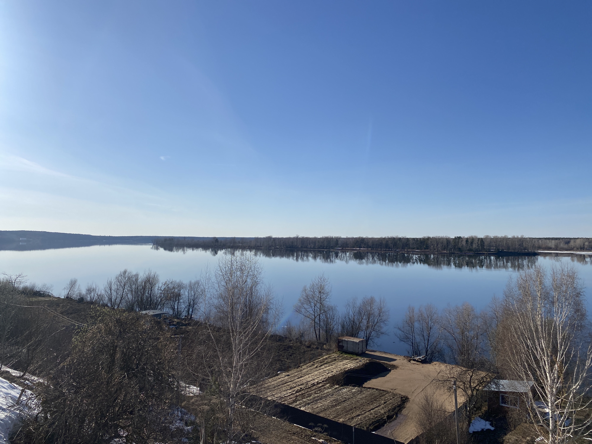 A serene, panoramic view captures a wide body of water, likely a large river or lake, under a vast, clear blue sky. The sun, visible as a bright flare in the upper left, illuminates the scene, suggesting a sunny day, possibly late winter or early spring.
The sky is a brilliant, unblemished blue with only the faintest wisp of clouds visible near the horizon. This clear sky is beautifully reflected in the calm, smooth surface of the water, creating a seamless transition from the sky to the distant horizon. On the far bank, a long, unbroken line of bare, dormant trees stretches across the frame, their silhouettes and reflections adding depth to the composition.
In the foreground, a sloped embankment leads down to the water's edge. This bank is dotted with numerous bare deciduous trees and tangled shrubs, indicative of the dormant season. Patches of lingering snow are visible on the ground, particularly on the left, confirming the seasonal transition. Evidence of human presence includes tilled garden plots with dark, exposed soil, suggesting preparation for planting. A small, weathered shed or container sits near the water on the right, and further right, partially obscured by trees, is another small building or house. A dirt path or road meanders along the bank, and a utility pole with wires is visible on the right.
The scene is devoid of any visible people or active events, conveying a sense of stillness and tranquility. No visible text is present in the image. The overall impression is one of quiet natural beauty, showcasing the subtle shifts of the seasons in a rural Russian landscape.