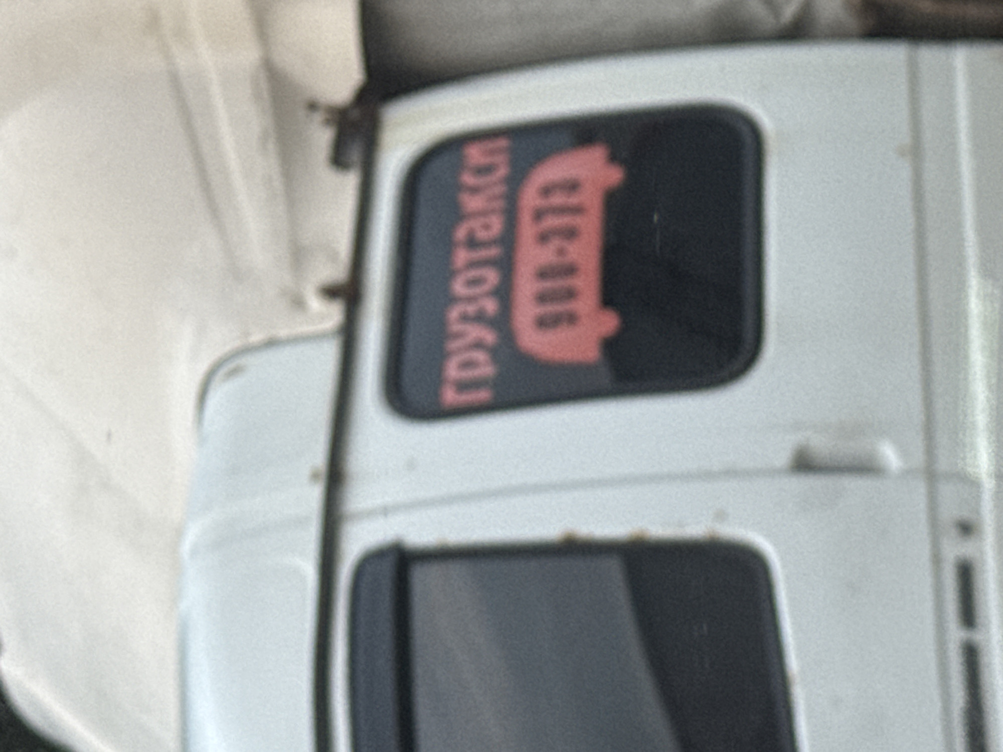 The image shows the side of a white delivery van parked on an overcast day. On the rear window, a red sticker displays the word "ГРУЗОТАКСИ" (GRUZOTAKSI), which translates to "freight taxi" or "delivery taxi" in Russian, above a red silhouette of a van and a phone number "908-373". The van appears to be parked on a street, though the surrounding environment is out of focus. The lighting suggests it is daytime, but the sky is heavily clouded. This imagery is consistent with a typical scene in Izhevsk, Russia, where such vehicles are commonly used for urban transport services.