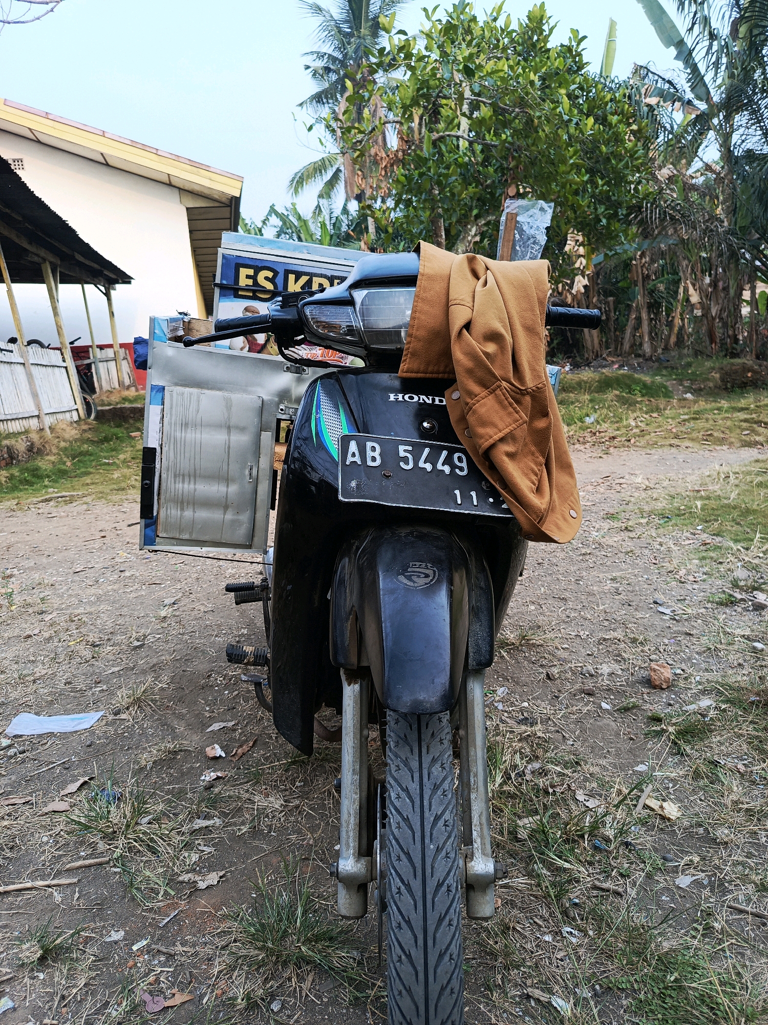 A motorcycle is parked on a dirt road in front of a white building with a yellow roof. The motorcycle has a brown jacket draped over its handlebars. A metal box, possibly for carrying goods, is attached to the side of the motorcycle. The background shows trees and vegetation, indicating a rural or semi-rural setting. The sky is clear and blue. The scene appears to be in Megang Sakti II, Indonesia, based on the motorcycle's license plate, which reads "AB 5449 11-20".