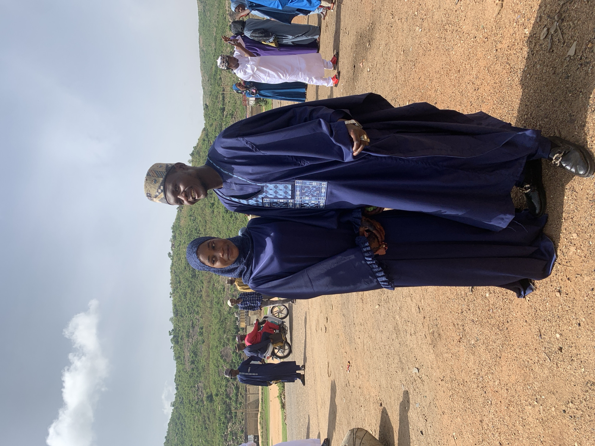 A man and a woman stand side-by-side, both dressed in navy blue traditional Nigerian attire, with a green, hilly landscape and a dusty tan-colored road in the background. The man wears a patterned cap and has a watch on his left wrist. The woman wears a head covering. They appear to be posing for a photo. Other people are visible in the background, some in colorful clothing and some standing on a motorbike. The location appears to be an open area in Naraguta Hausawa, Nigeria.