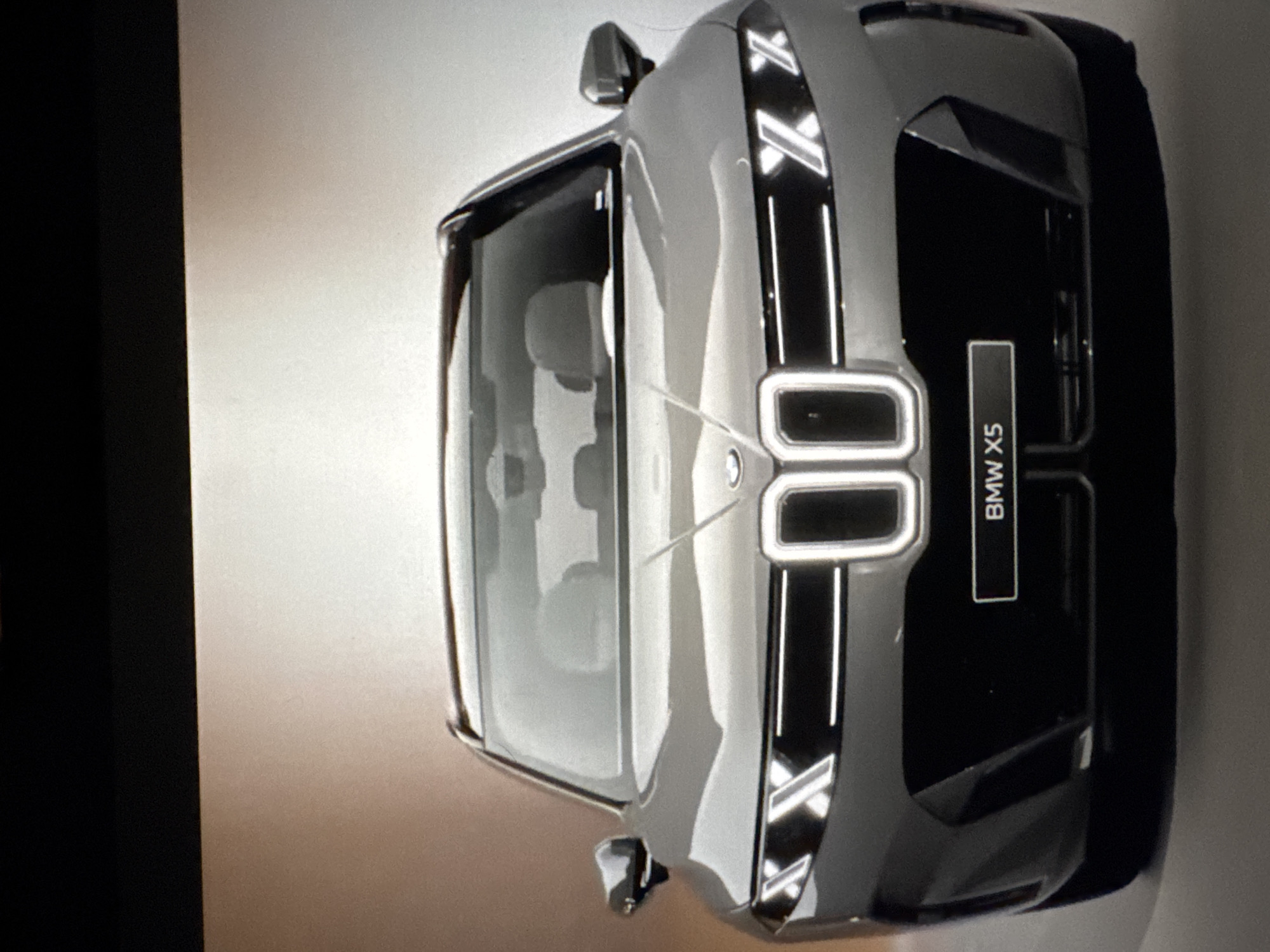 The image shows a frontal view of a grey BMW X5. The car has a distinctive grille with illuminated vertical elements, and sharp, angular headlights. A black license plate with "BMW X5" written in white is visible below the grille. The car is positioned against a gradient background that transitions from a darker hue at the top to a lighter, almost white color at the bottom. The lighting suggests a studio setting or a controlled environment, with the car being the sole focus. There are no people or any urban environmental elements visible in the image, therefore it is impossible to determine its location in Sant'Angelo di Piove di Sacco, Italy.